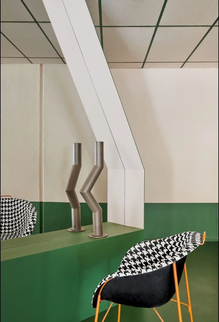 Interior space with green counter, black and white patterned chair, two metallic tube vents, and a white angular structure against a beige wall with green lower section. Suspended ceiling with square tiles.