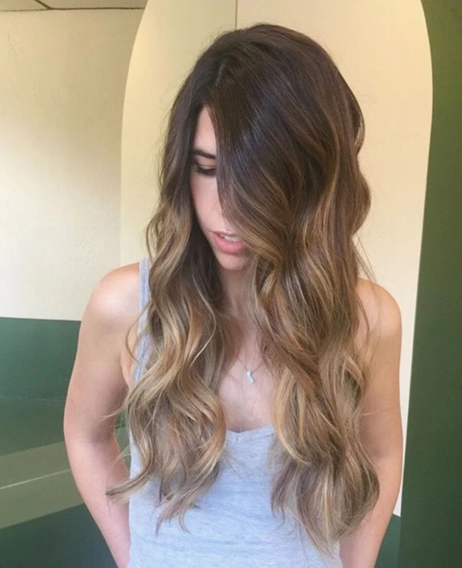 Young woman with long, wavy, brown blonde ombre hair looking down, wearing a gray tank top, in front of a green and beige background.