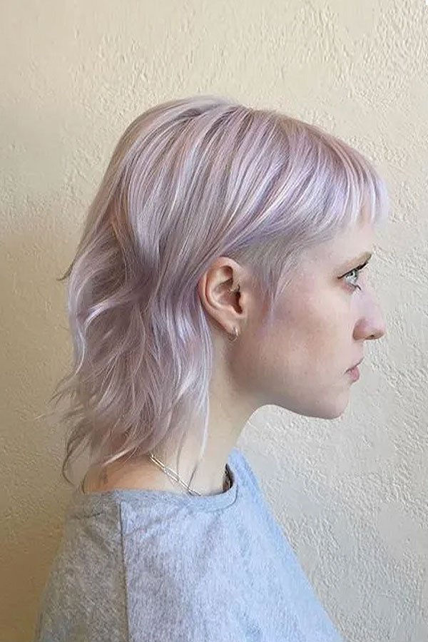 Profile of a woman with short, wavy, pastel purple hair standing against a beige wall.