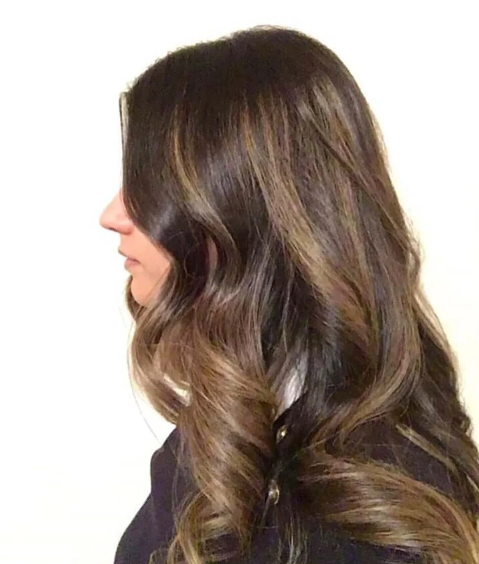 Profile side view of a woman with long, wavy brown hair and soft highlights, dressed in a black top, against a plain white background.