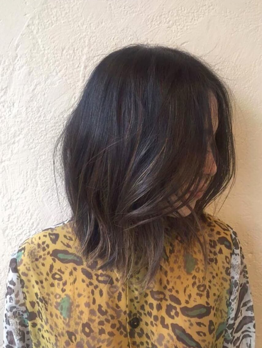 A woman with shoulder-length dark hair with light highlights, wearing a yellow and brown leopard print shirt, standing against a textured beige wall.