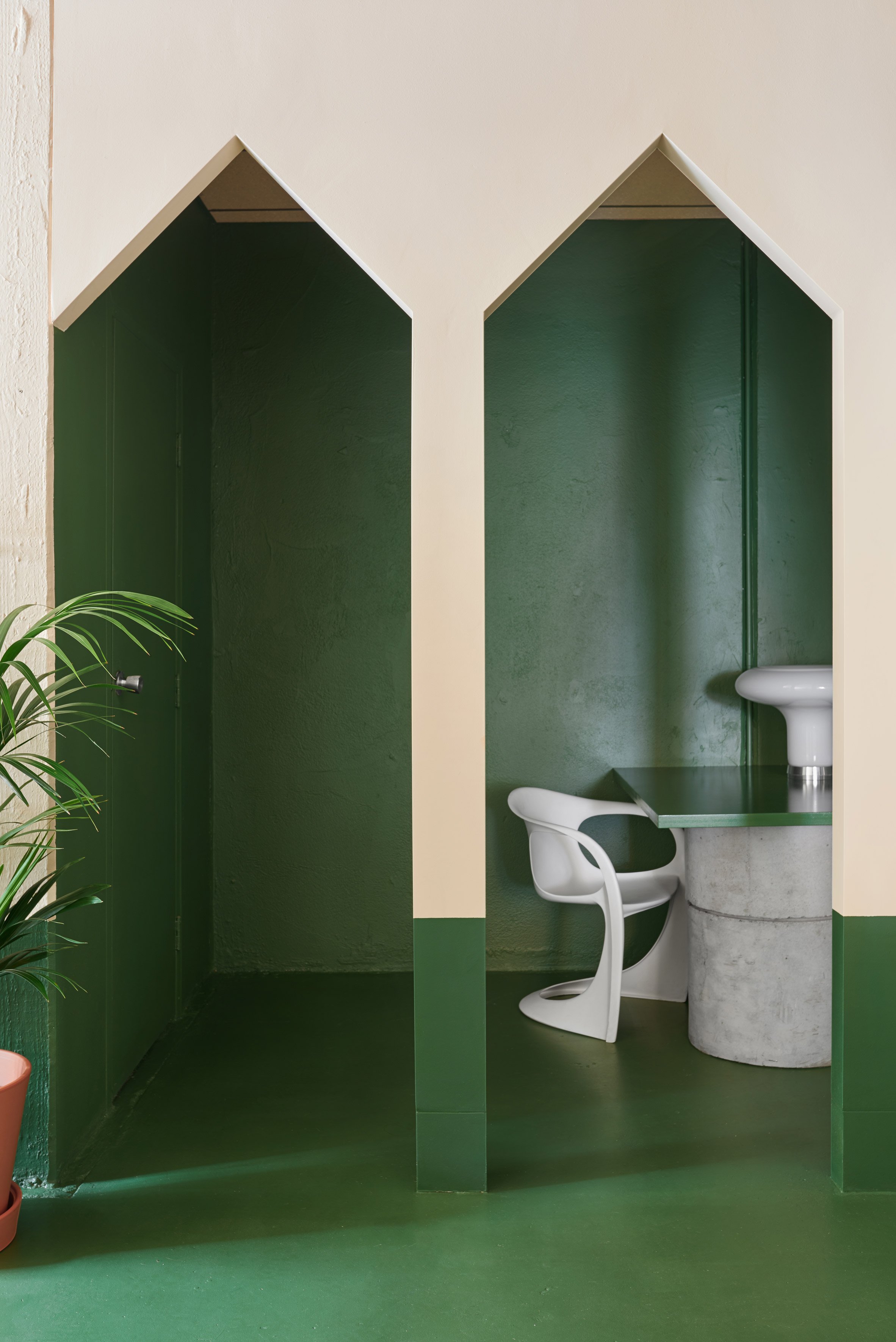 A minimalist bathroom corner with a green and white color scheme, a white chair, a small green table, and a white pedestal sink, framed by a white wall with green accents.