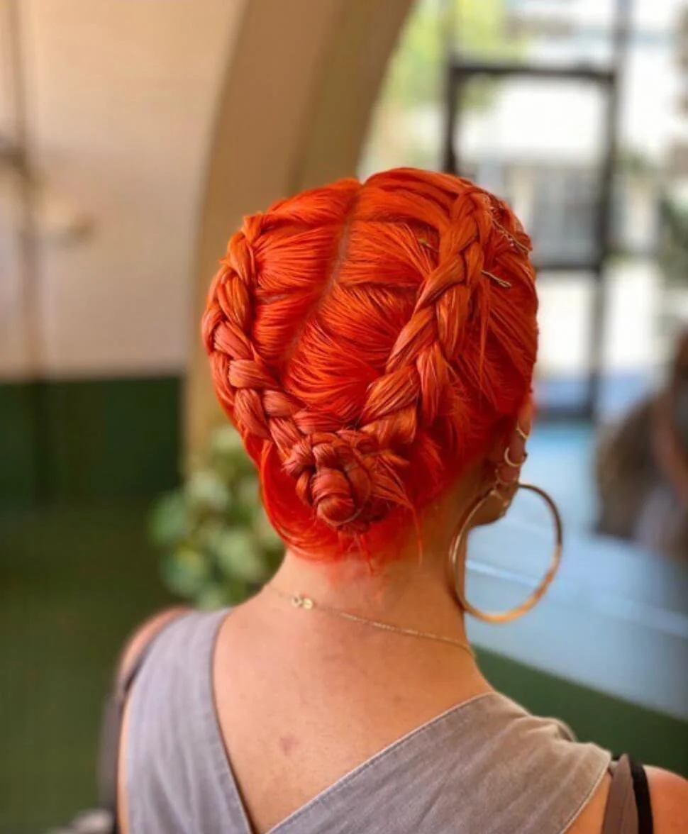 A person with vibrant red braided hair styled in an intricate updo, wearing large hoop earrings and a thin necklace, seen from the side inside a room with a window and plants in the background.