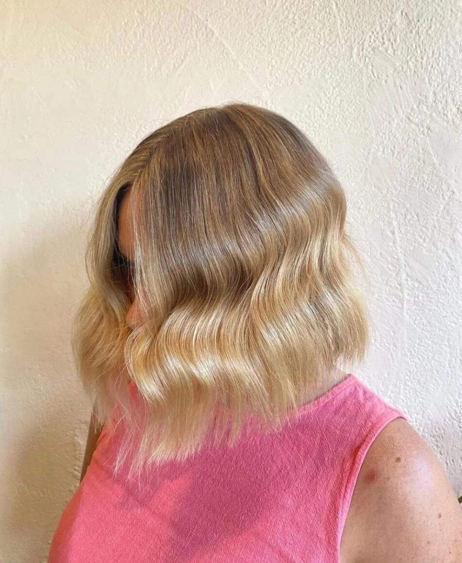 Woman with shoulder-length wavy blonde hair, wearing a pink sleeveless top, standing against a textured beige wall.