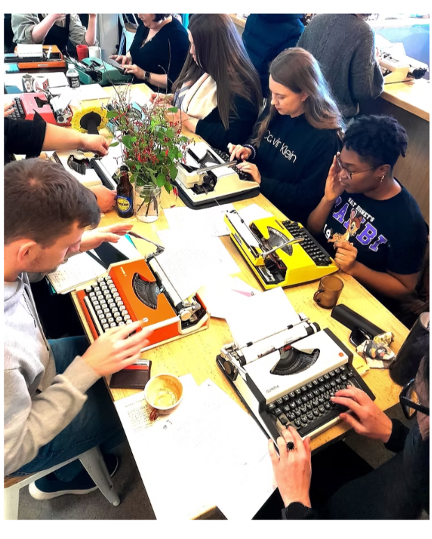 Group of people sitting around a table using vintage typewriters, with papers, a potted plant, drinks, and personal items on the table.