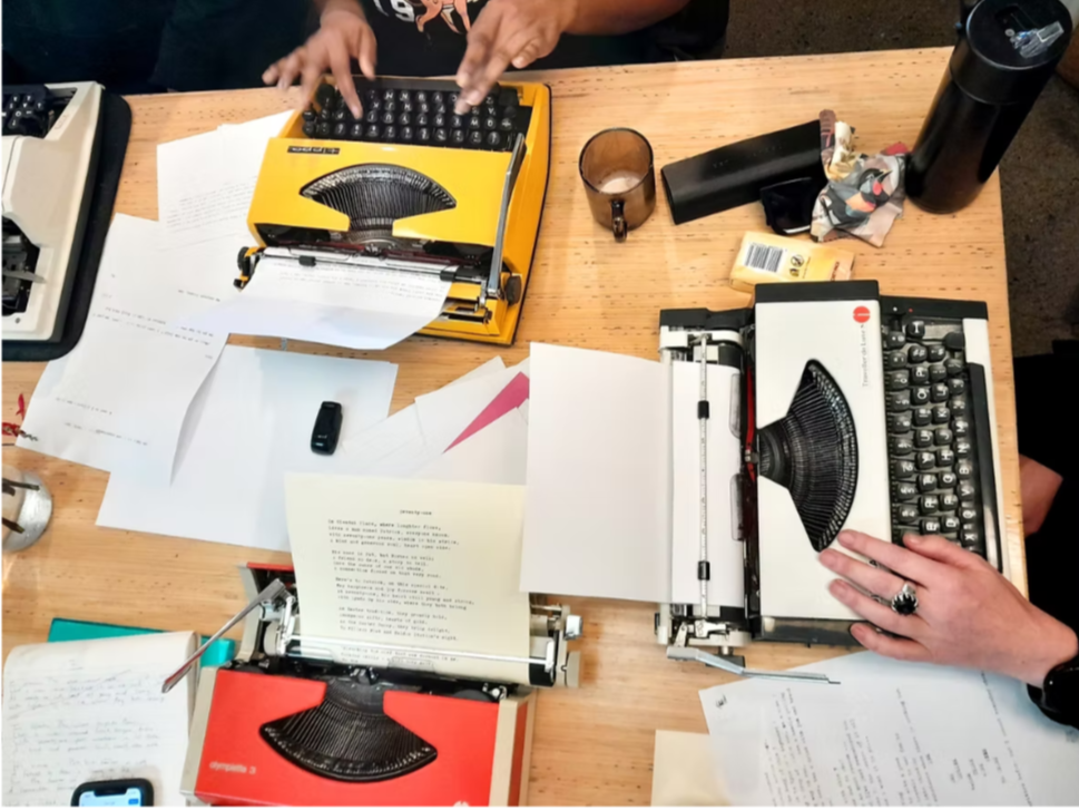 Two vintage typewriters on a cluttered wooden table, surrounded by papers, a remote control, a black thermos, a glass, and a hand with rings pressing keys on the right typewriter.
