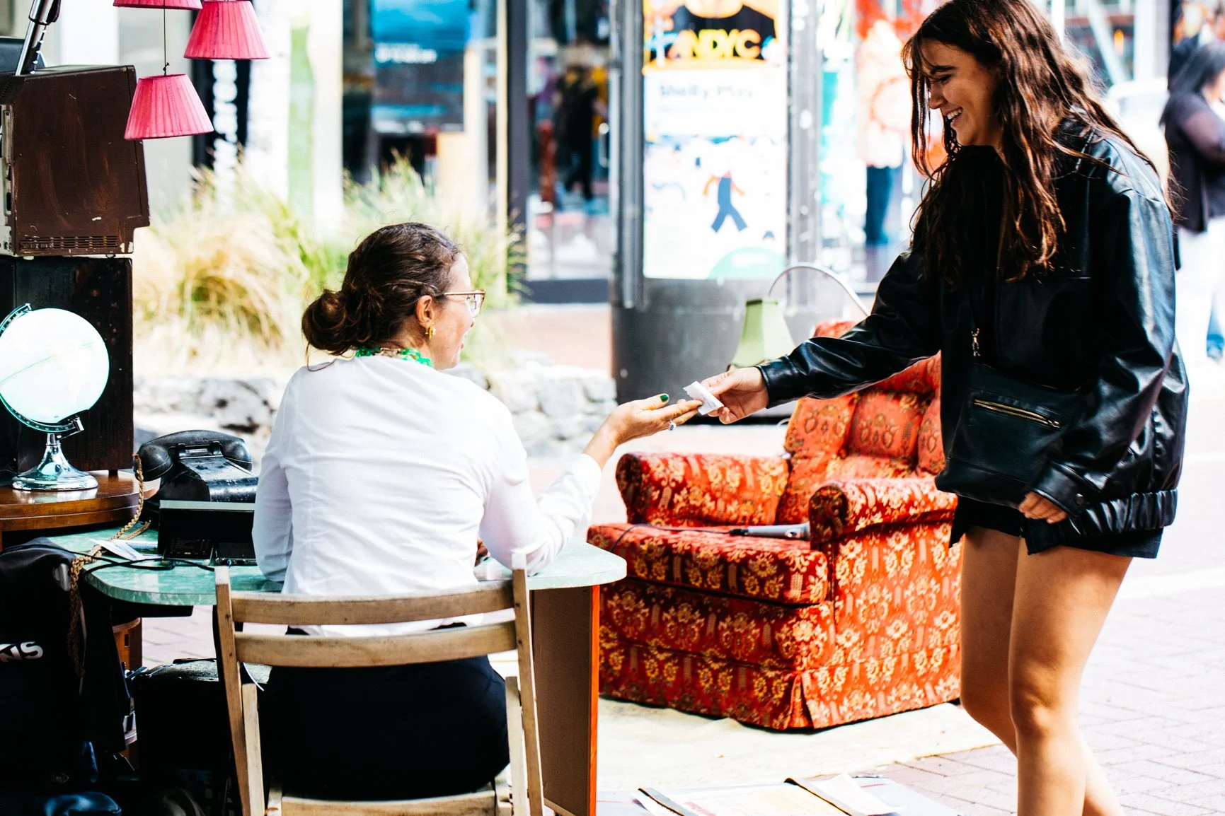 A woman in a black leather jacket hands a folded bill to a seated woman with glasses and a white blouse, sitting at a small outdoor table with a vintage-style colorful armchair nearby. The setting appears to be a street scene with pedestrians in the 