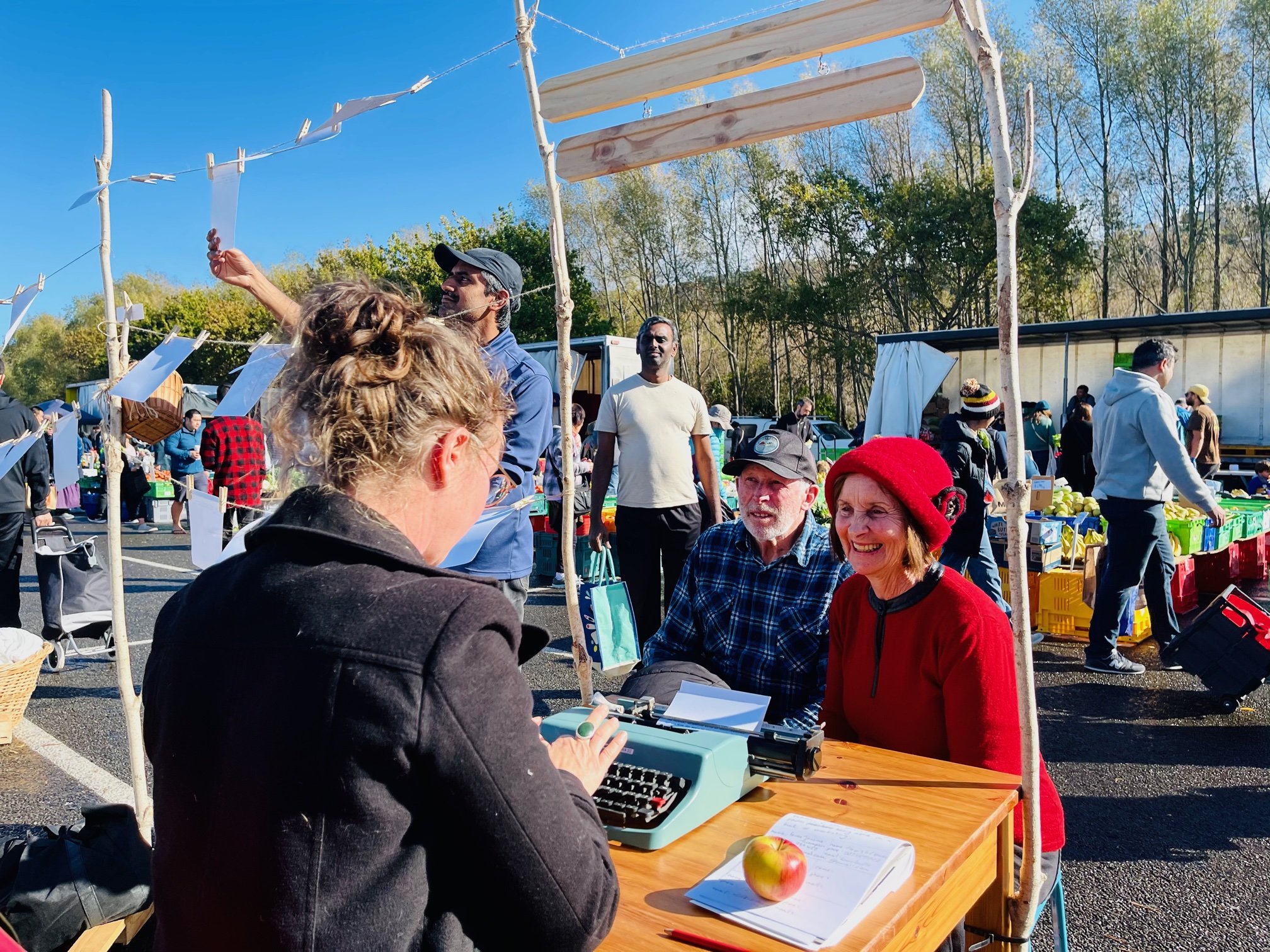 People gather at an outdoor market on a sunny day, with some engaged in conversation and others shopping for produce. There are tables filled with apples and other goods, and trees with green leaves in the background.