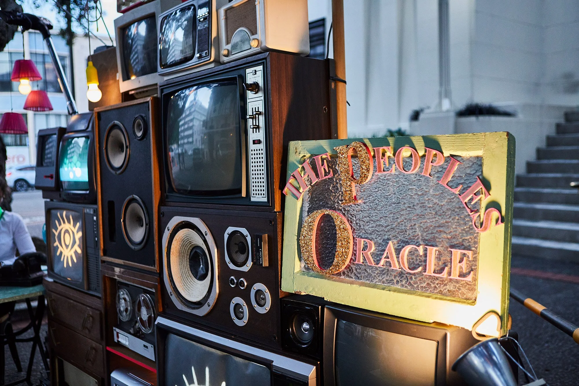 A display of vintage televisions, radios, and speakers arranged on a wooden stand at an outdoor event. There is a colorful sign that reads 'The Olde Laffle' and 'ORACLE' decorated with glitter nearby.