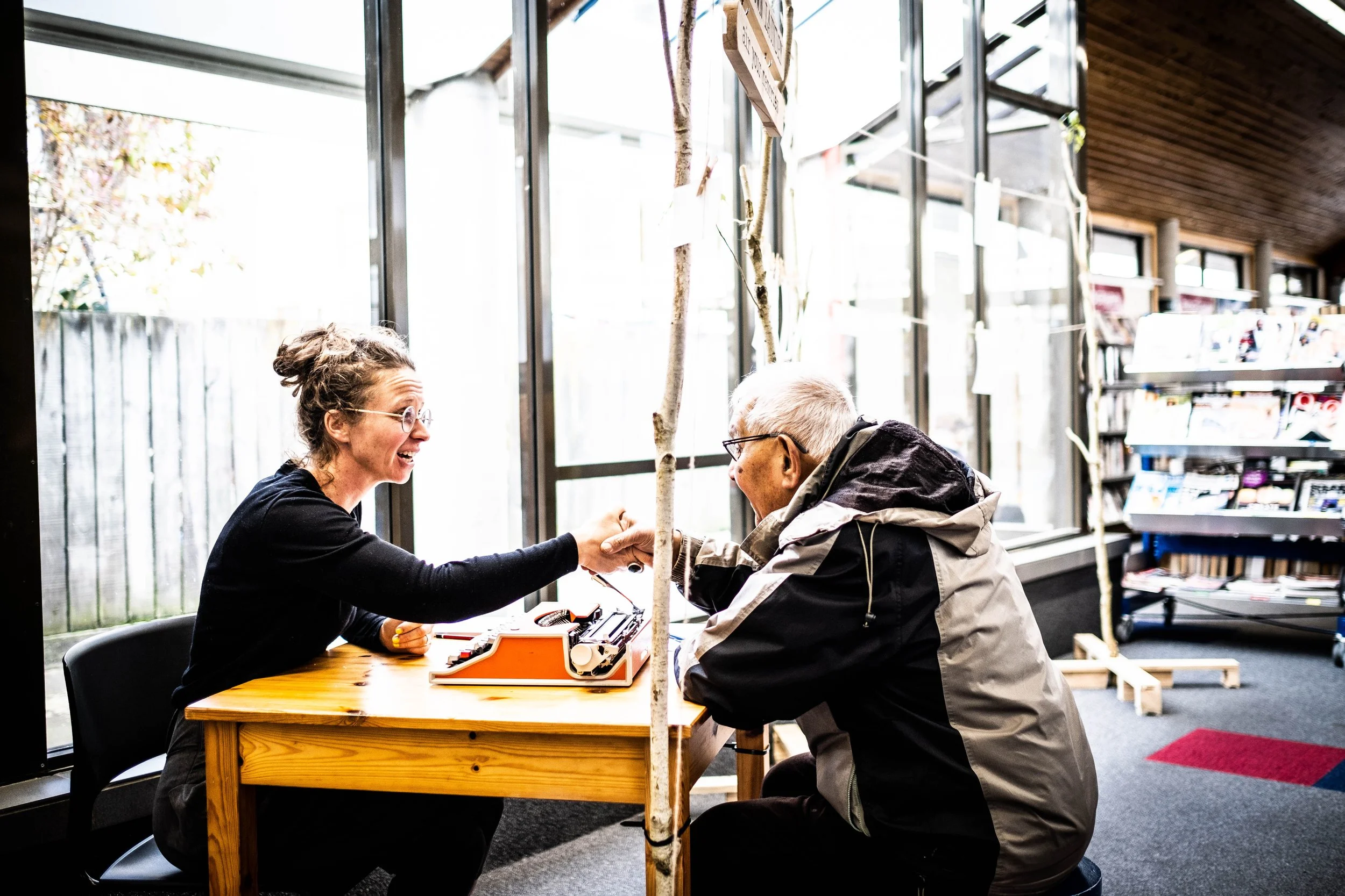 Two people, a young woman and an elderly man, sit at a wooden table engaged in an activity involving small tools and a device. They are indoors near large windows with a tree outside and shelves filled with books inside.