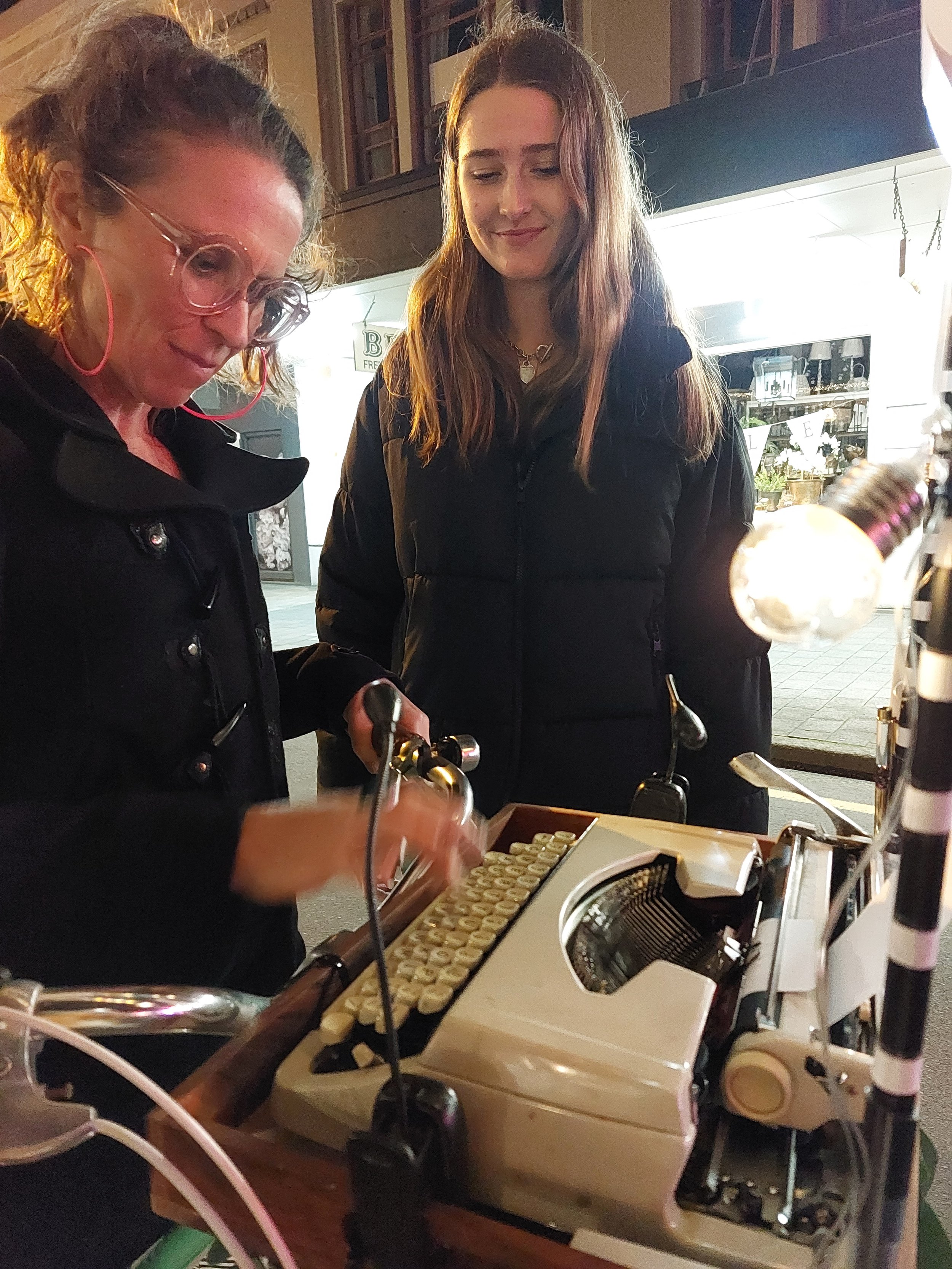 Two women standing at a street food stall at night. One woman is operating an electric grill with small food pieces, and the other woman is watching. The background has a store window decorated with a bunting and some display items.