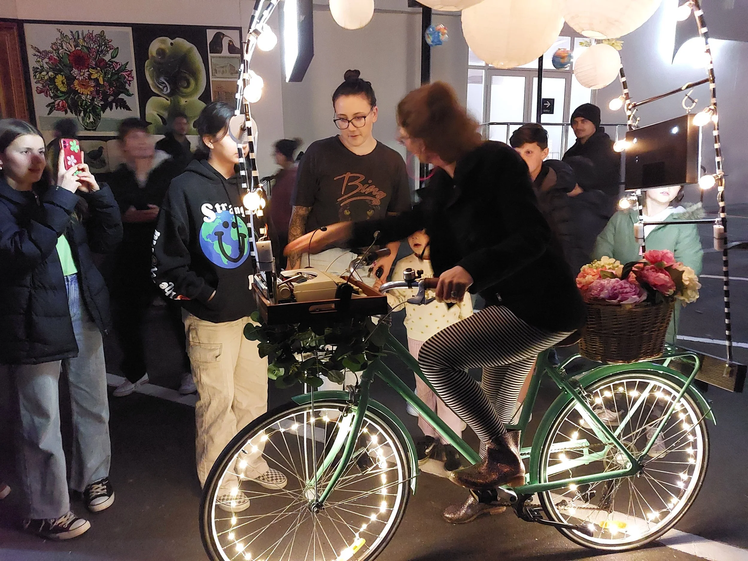 A group of people gathered around a vintage bicycle decorated with string lights, with a woman sitting on the bike, inside a location decorated with hanging paper lanterns and wall art.
