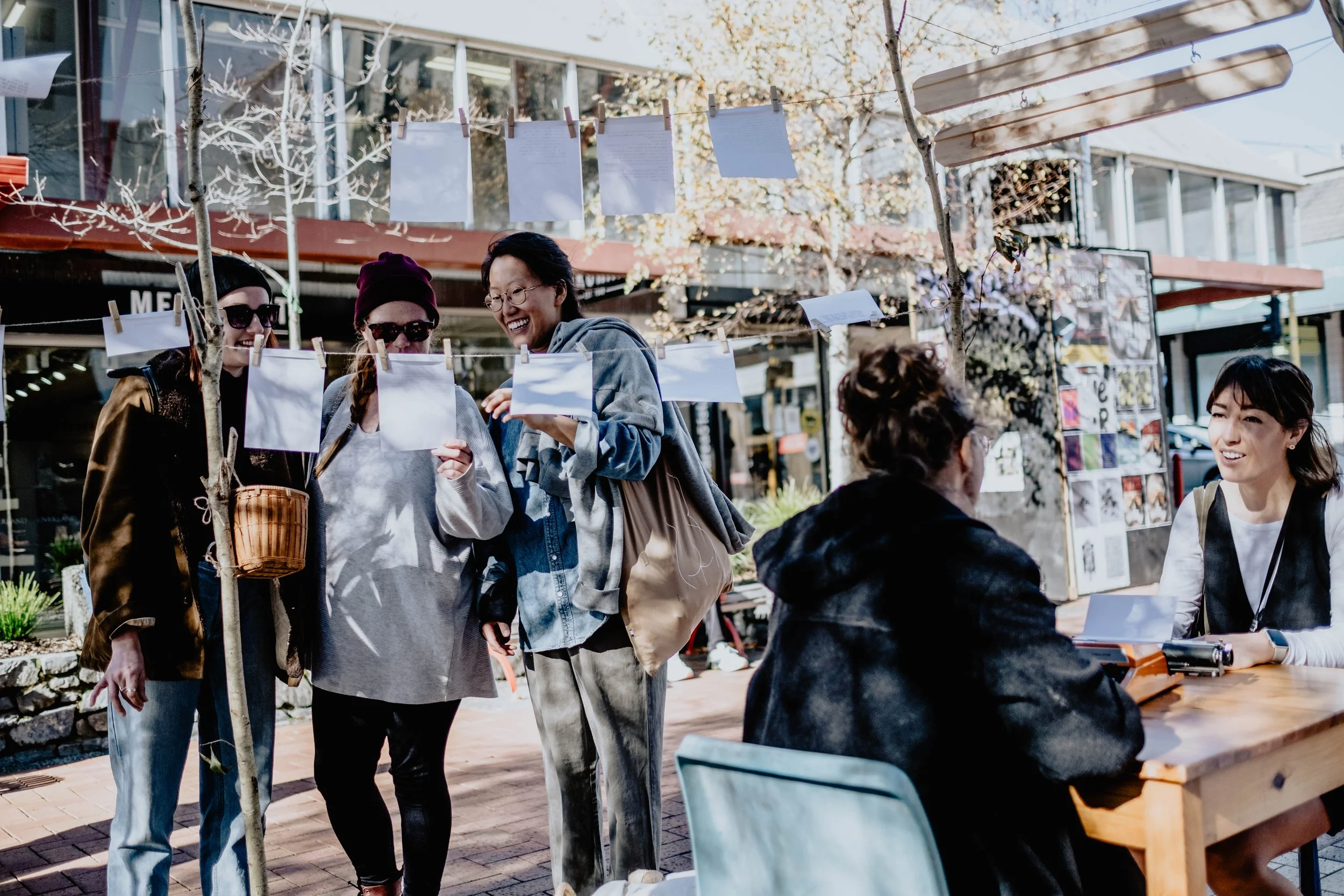 Group of people conversing at an outdoor market stall with white papers hanging on strings and trees in the background.