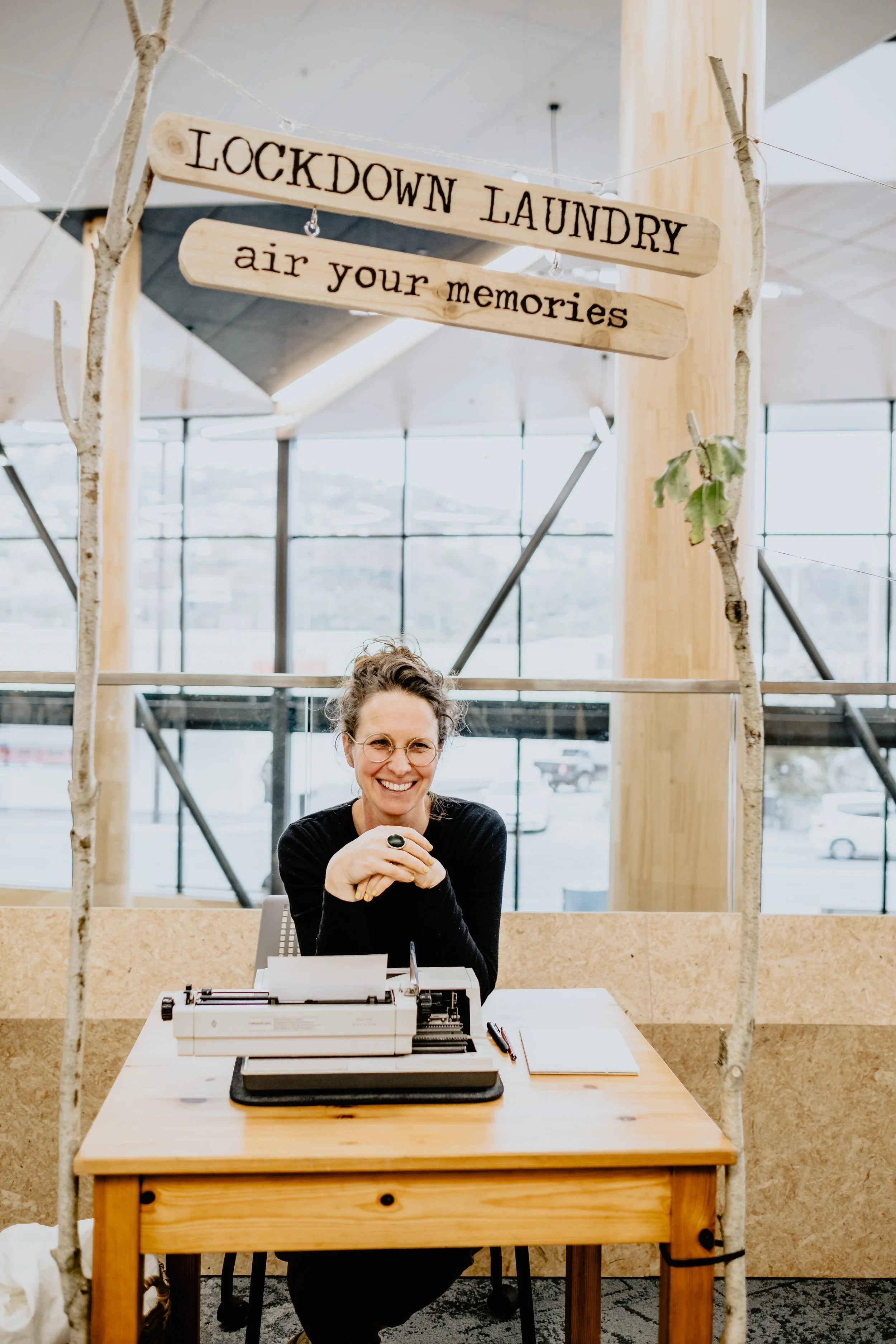A woman with glasses and curly hair smiling at a table with a typewriter and papers in an office or lobby with large windows and a sign that reads 'Lockdown Laundry, air your memories.'
