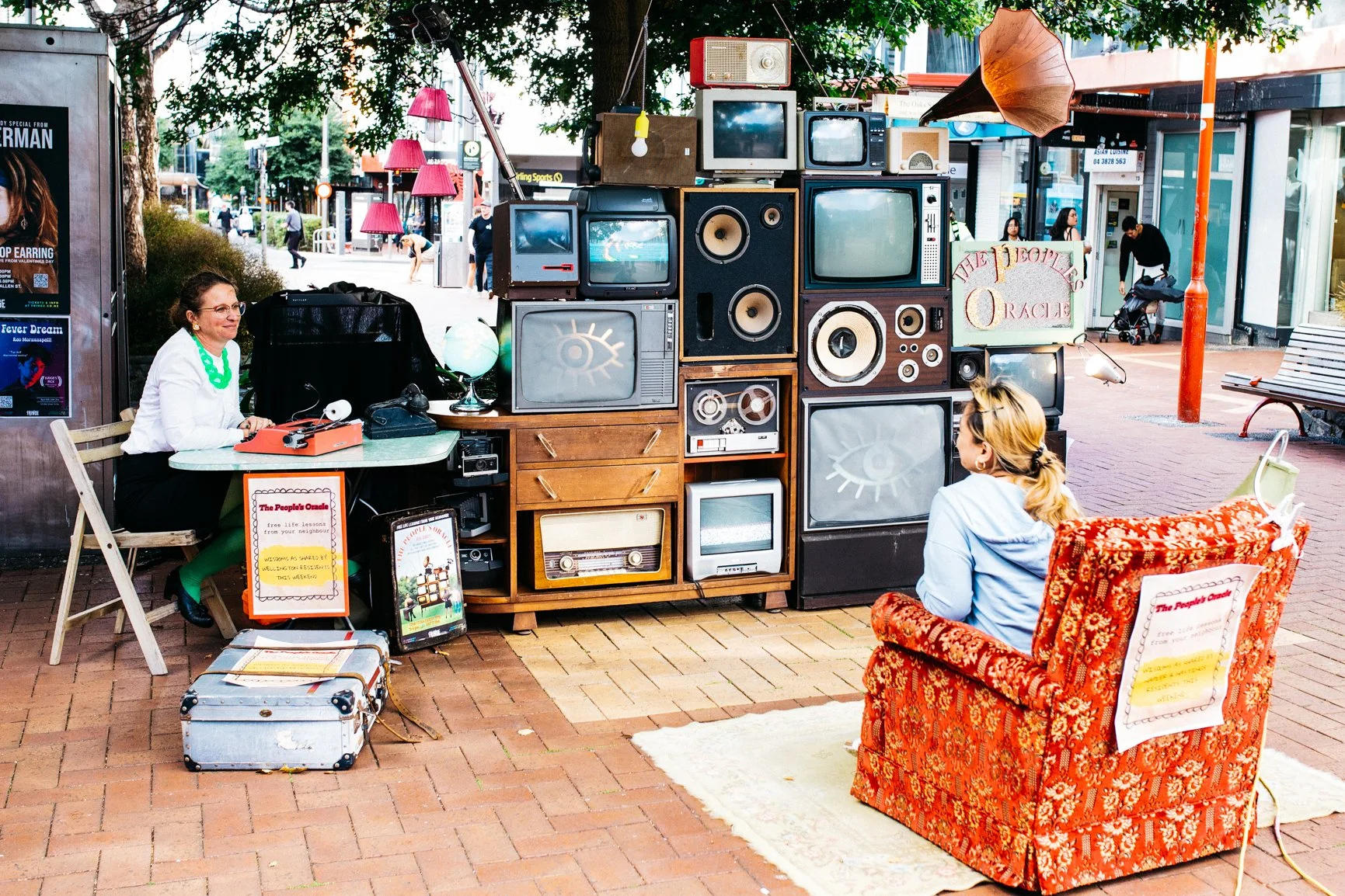 Outdoor street scene featuring a vintage display of old televisions and radios stacked in a creative arrangement. There are two women sitting and conversing near the display, one on a wooden chair and the other on a patterned upholstered armchair. Th