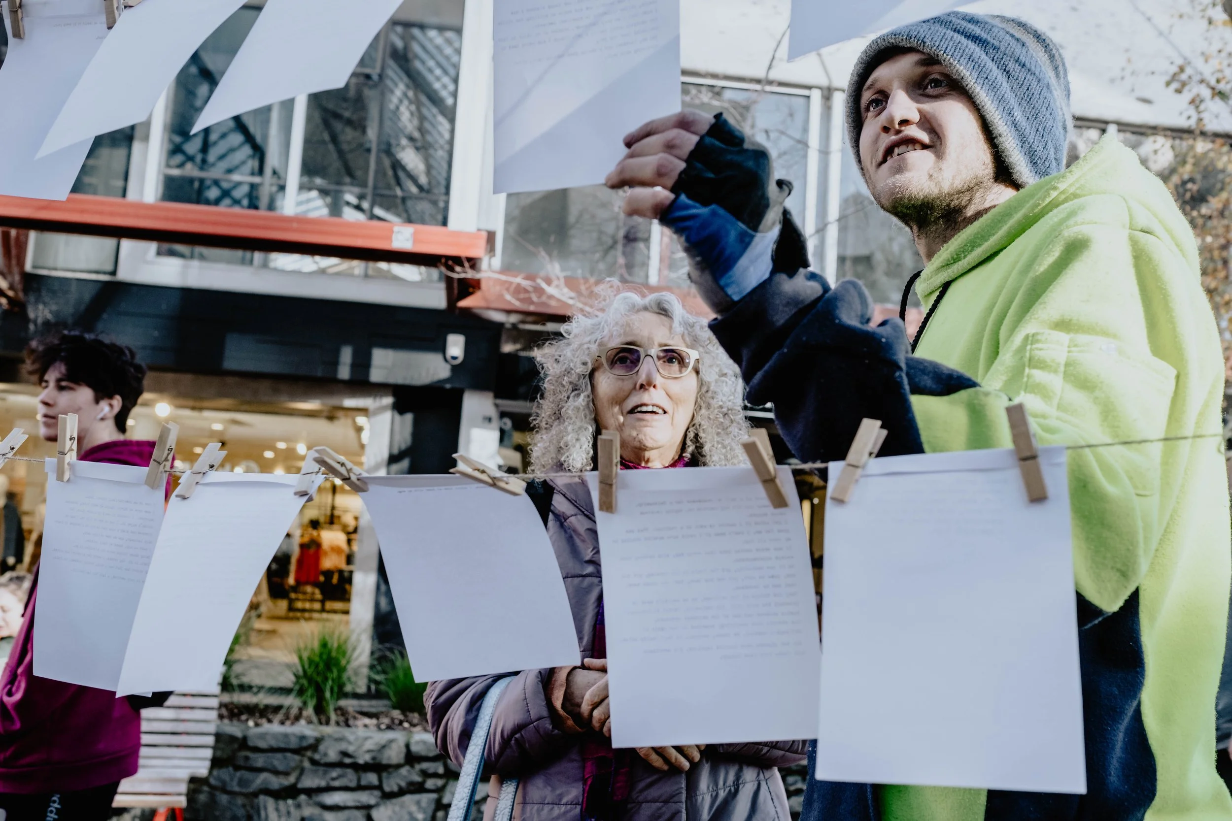 People hanging papers on a clothesline outside, with a woman wearing glasses and a gray jacket, an older woman with curly gray hair, and a young man in a green hoodie, with a background of buildings and trees.