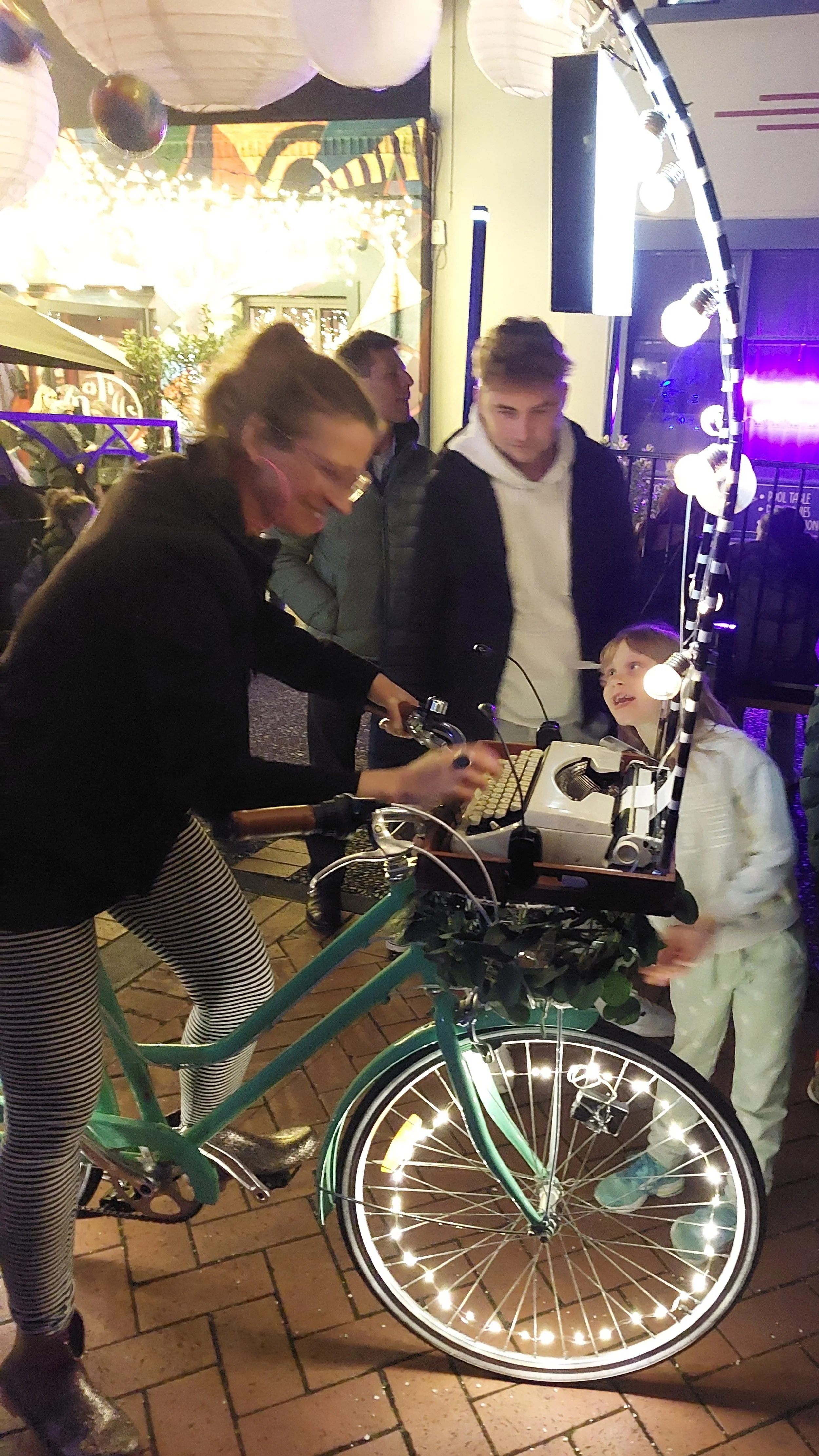 A woman riding a bicycle adorned with small lights, with a group of people gathered around her at night. The bicycle's front wheel is illuminated by lights, and there are large paper lanterns and balloons hanging overhead at an outdoor event.
