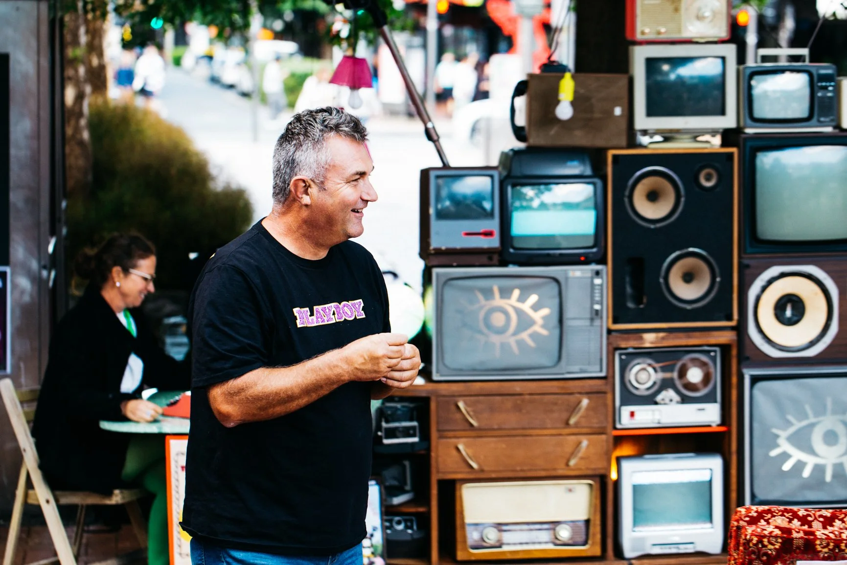A man with gray hair smiling and wearing a black T-shirt with 'PLAYBOY' written on it, standing in front of a display of vintage televisions and speakers at an outdoor market. A woman is in the background writing at a booth.