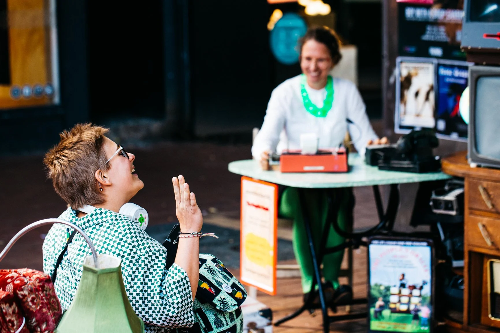 A woman with short hair and glasses, wearing a green patterned dress and headphones around her neck, appears to be happy and waving her hand while sitting at a table. Behind her, a smiling woman dressed in white, wearing a bright green necklace, is s