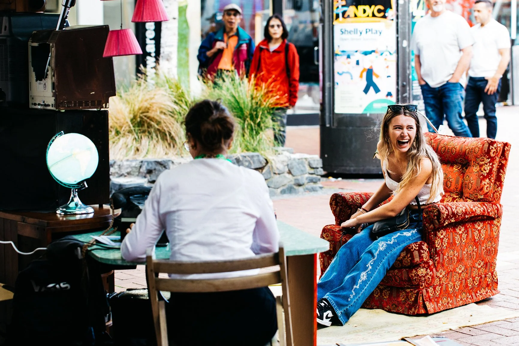 A woman with long blonde hair laughing and sitting on a vintage red patterned armchair outside on a city sidewalk. She is wearing a white top and blue jeans with embroidery, sunglasses on her head. A person with short hair, seen from behind, is sitting at a table facing her inside an outdoor cafe, with a vintage TV and globe on the table. People are walking in the background near a storefront with a sign that reads "Shelly Play."