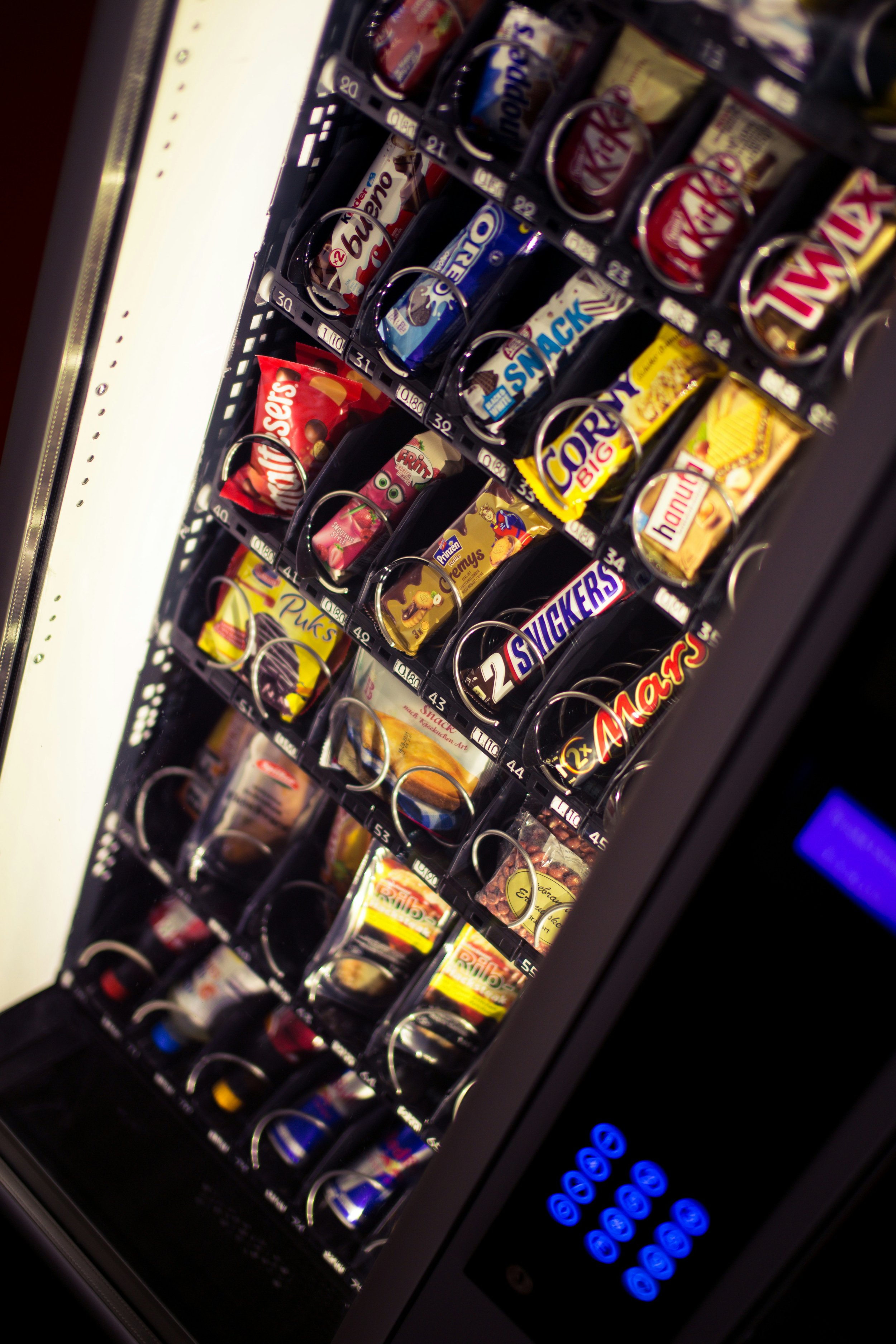 View of a vending machine with various snack items including candy bars and chips, illuminated by bright lights, with a digital payment keypad on the right side.