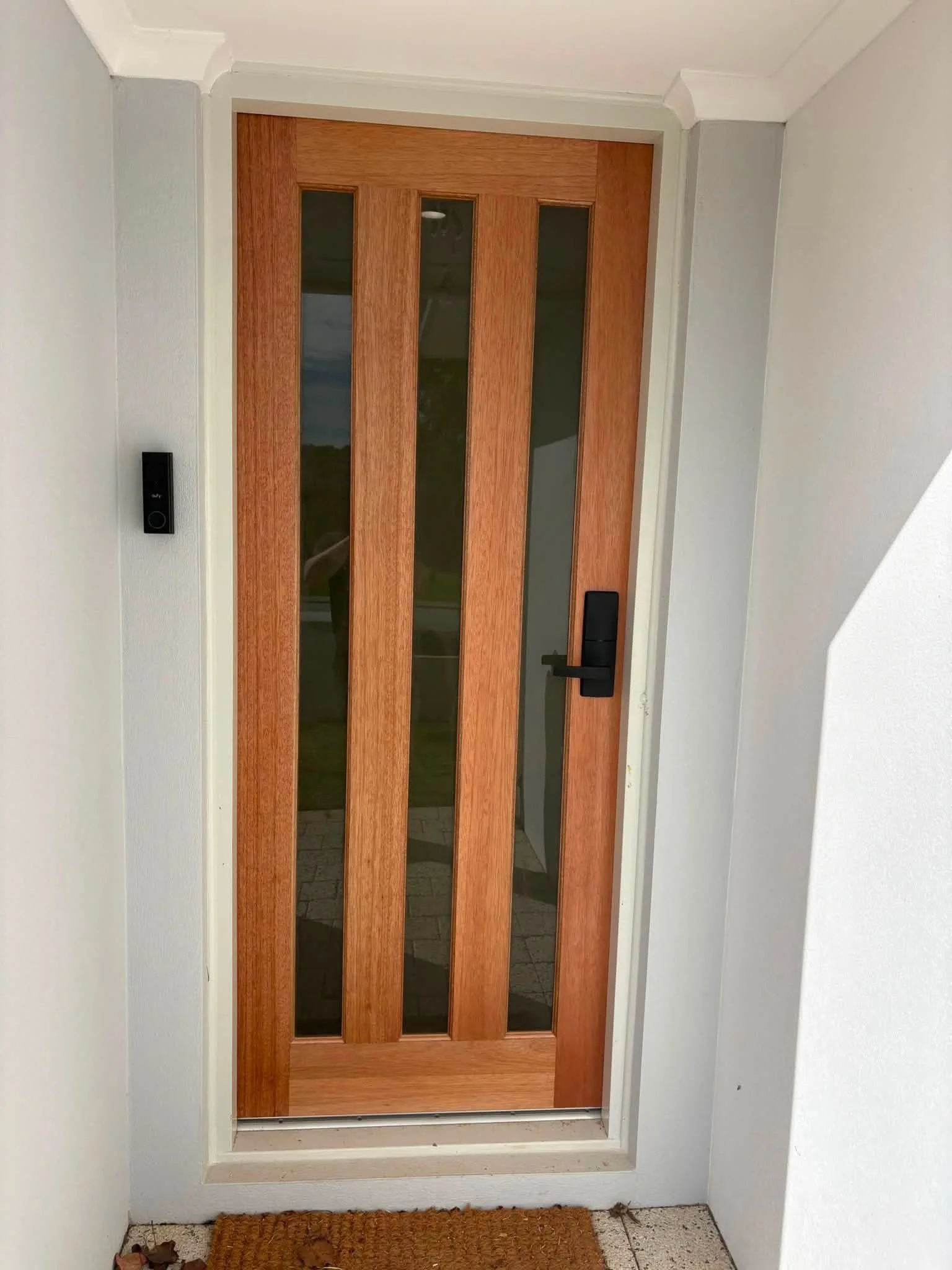 Front door with vertical wooden panels and glass inserts, black handle and lock, in white wall, with a doormat and some fallen leaves inside doorway.