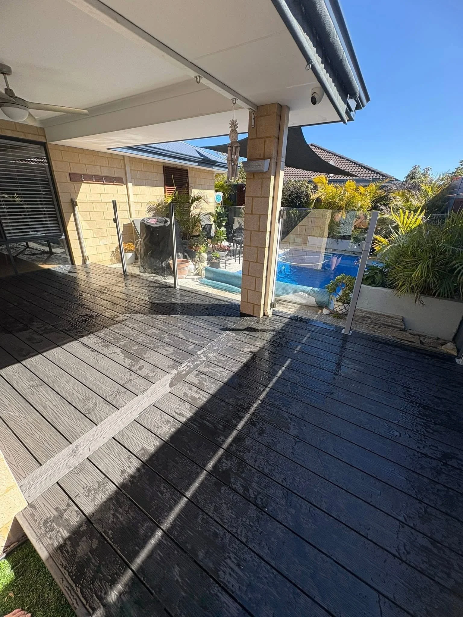 View of a backyard patio with a wooden deck, glass fence panels, potted plants, and a swimming pool in the background. The sky is clear and blue.