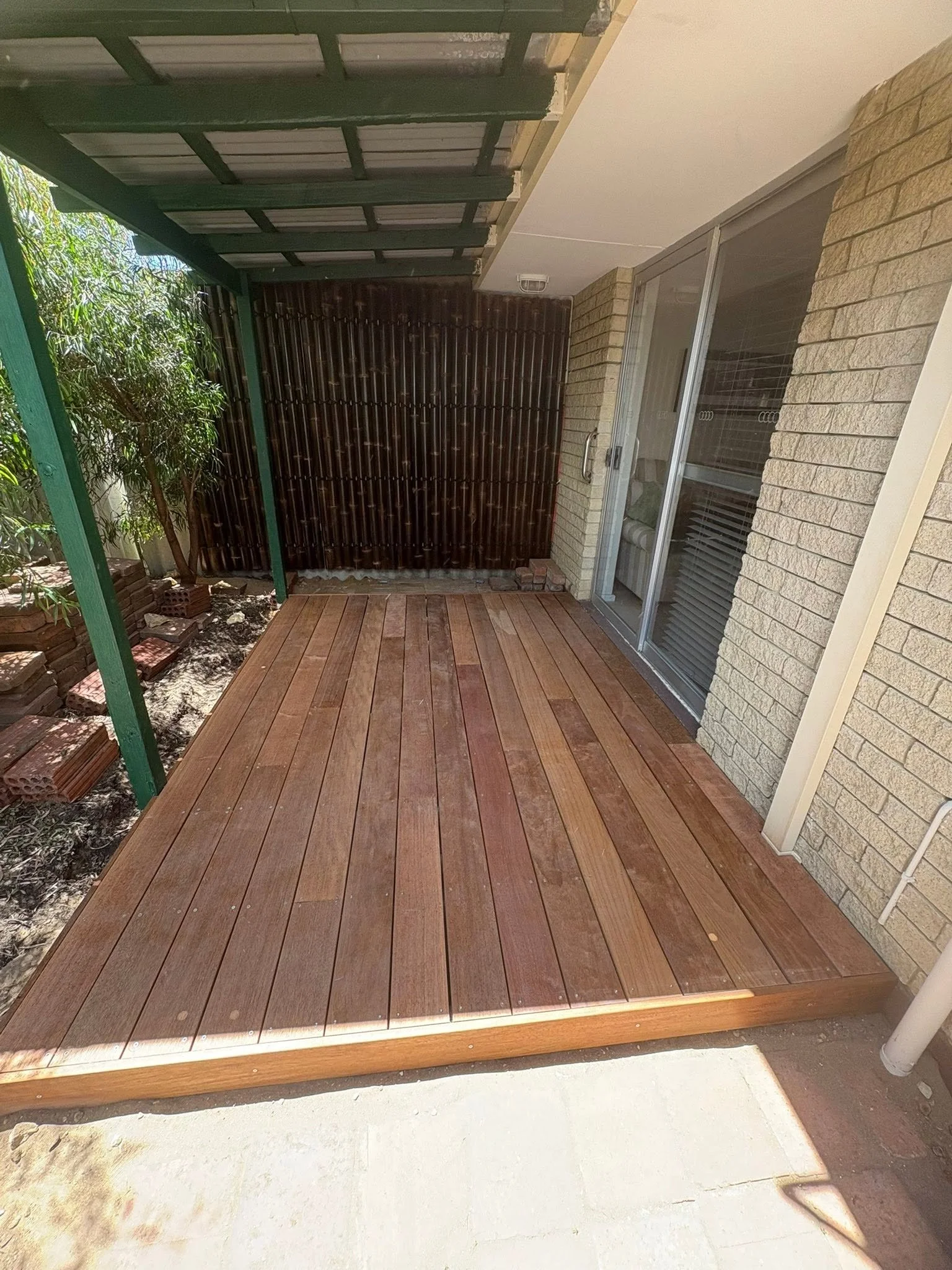 A newly installed wooden deck with a brick border, adjacent to a brick house with glass sliding doors, partly covered by a green metal pergola. There is a small garden area with plants and bricks on the left side.