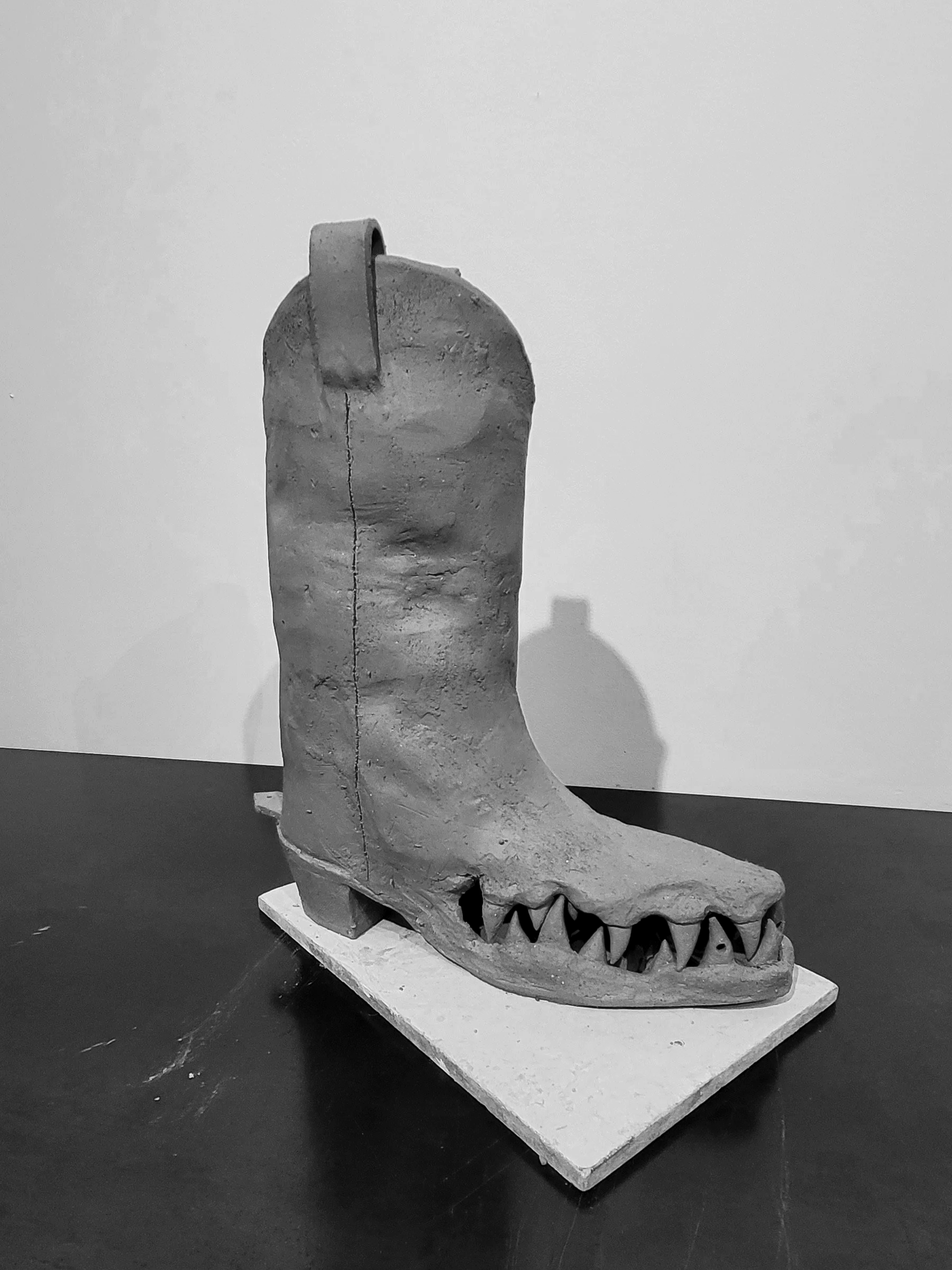 A ceramic cowboy boot in the process of being made in a pottery studio, displayed against a plain background.