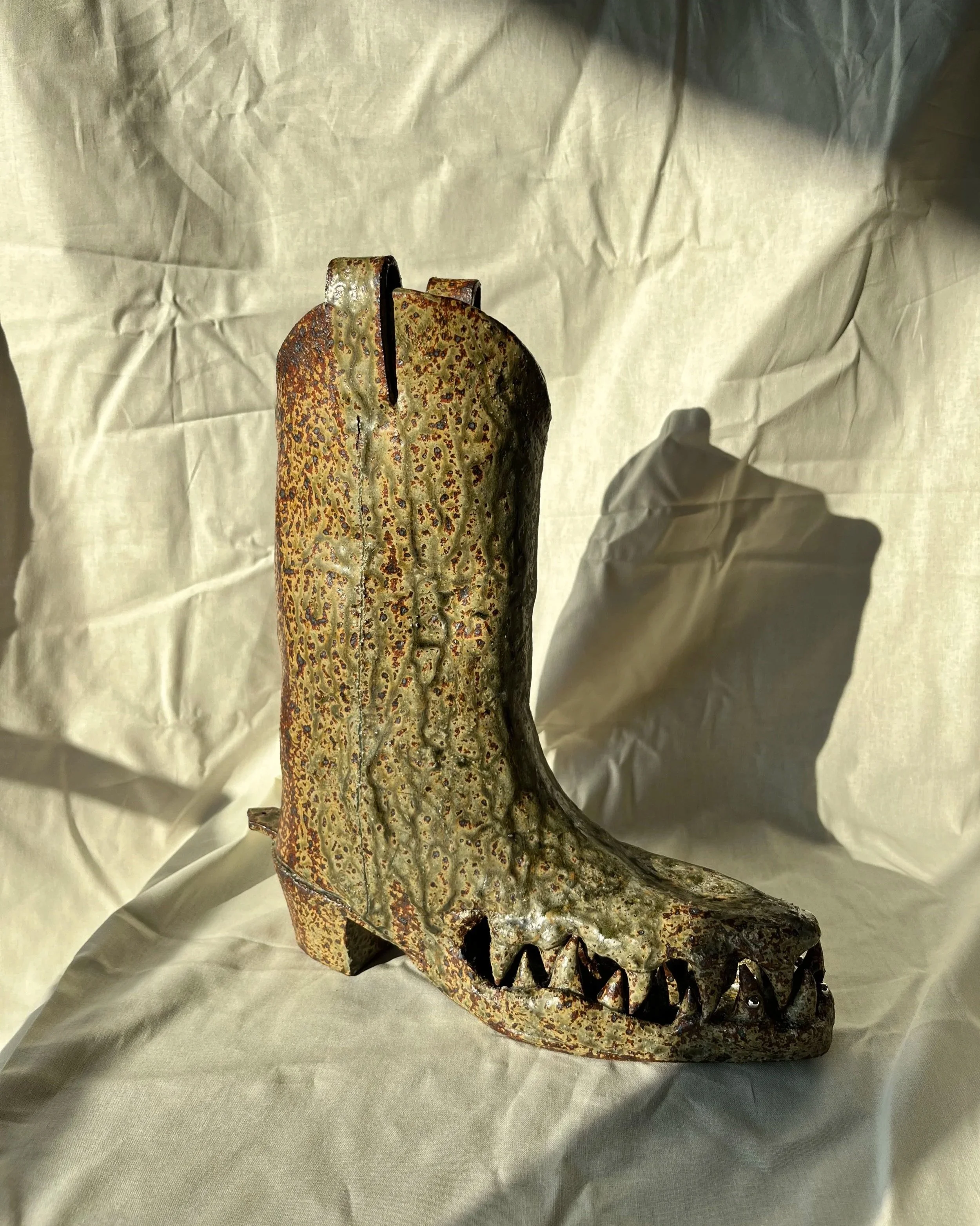 A ceramic cowboy boot with a crocodile jaw as the boot foot, with a green and rust coloured glaze set on a light coloured fabric background.
