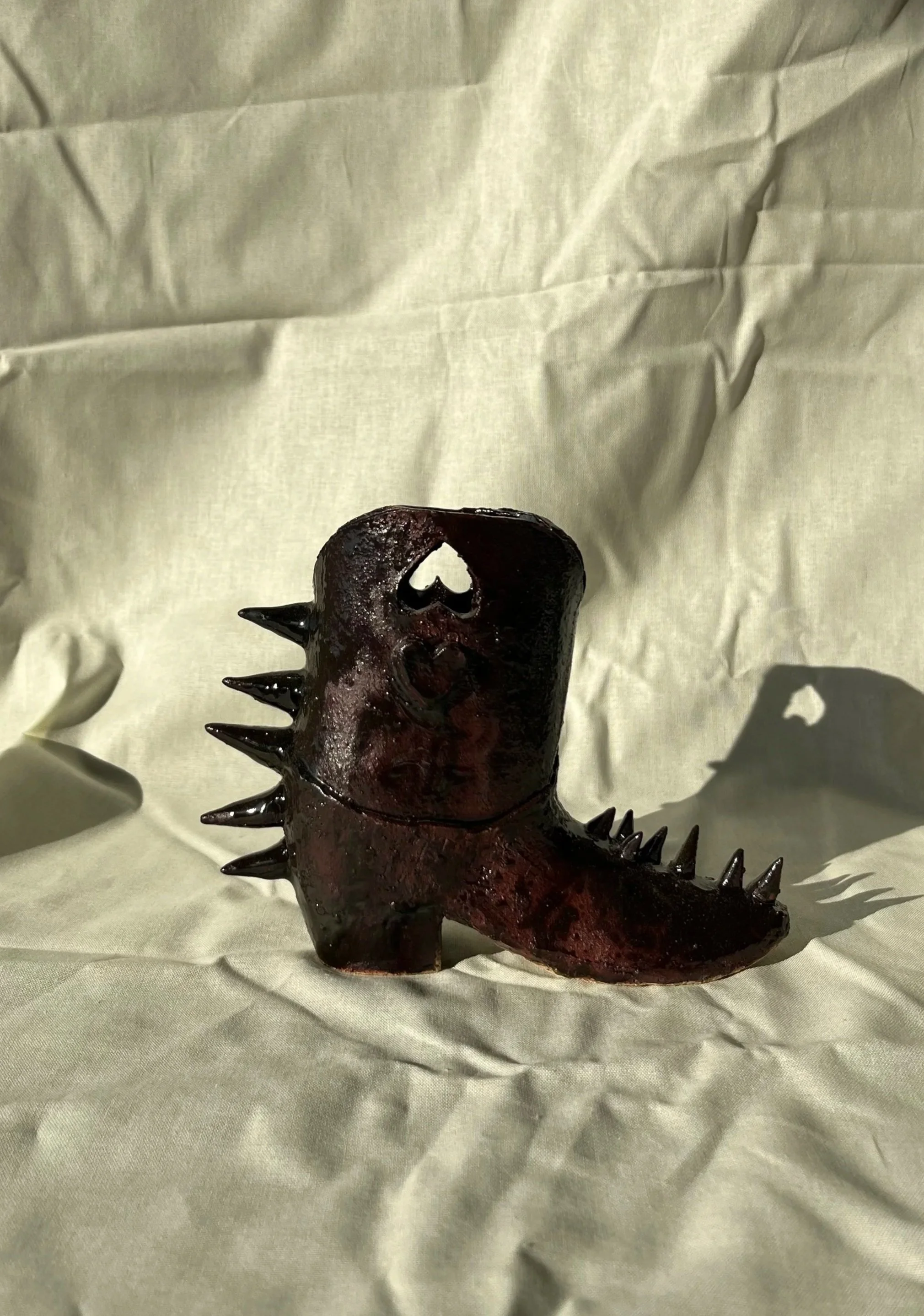A mini ceramic cowboy boot with a dark, glossy glaze and a heart-shaped cutout, sitting on a wrinkled, light coloured fabric background.