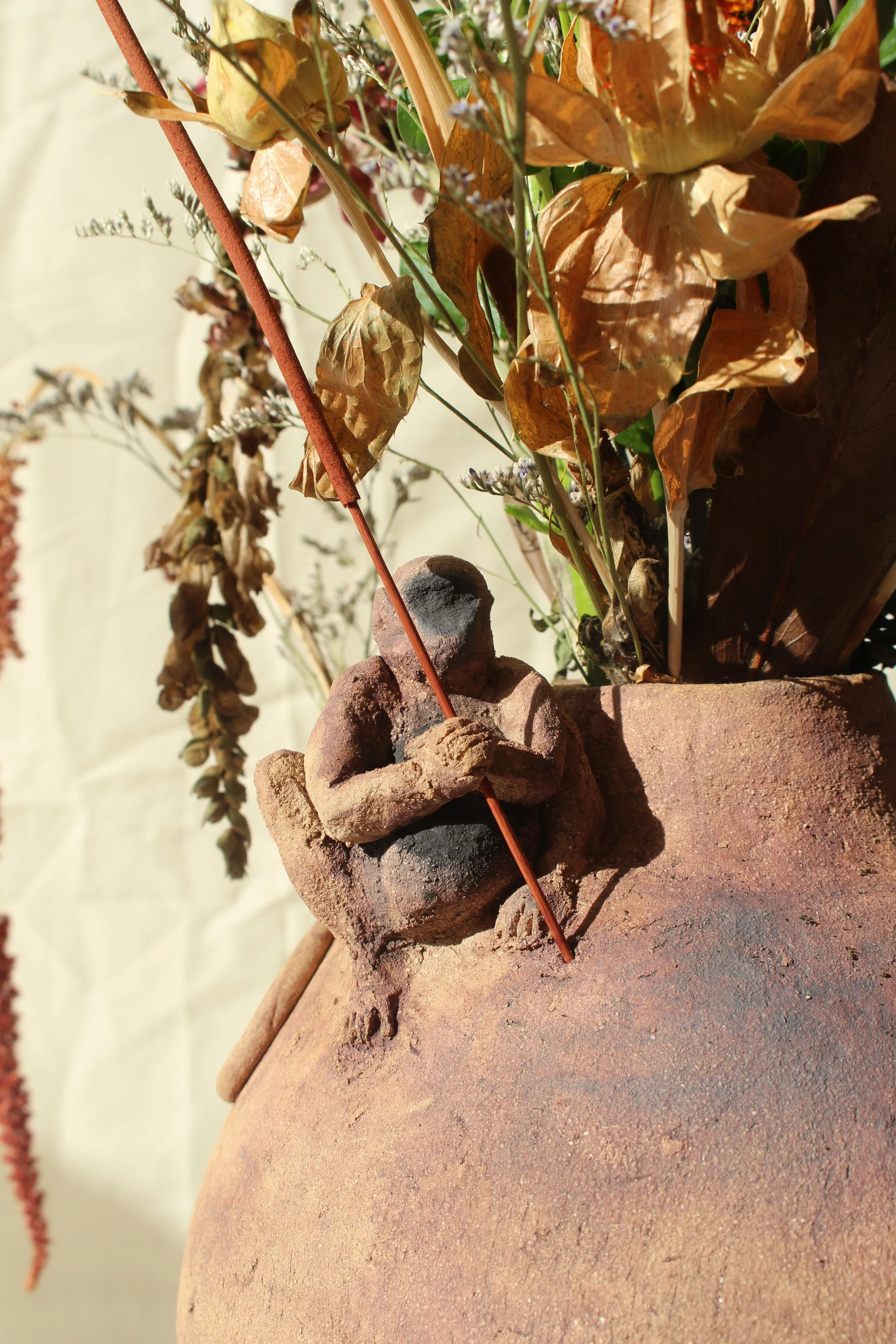 Close-up of a ceramic vase with little monkey sitting on it holding an insent stick. The vase holds dried leaves and flowers. The sculpture is on a light coloured surface.