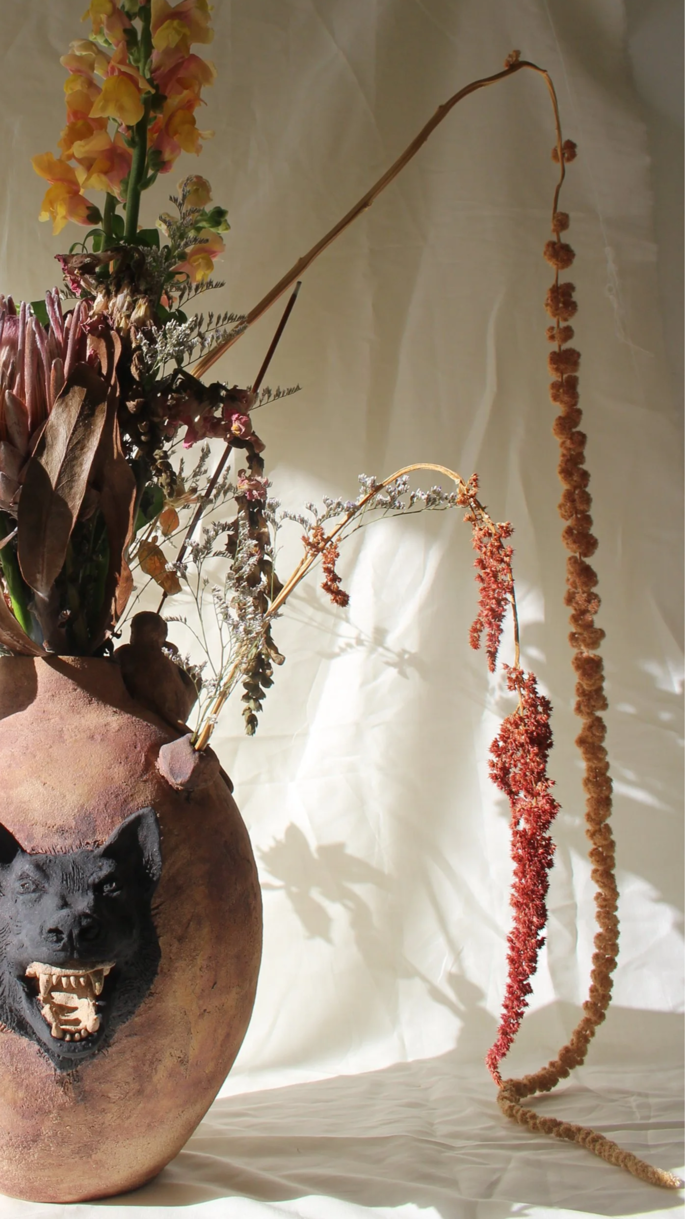 A ceramic vase with dried and fresh flowers and foliage, the vase has a black wolf snarling on the front. It is pictured on a white fabric backdrop with hanging dried flower stems.