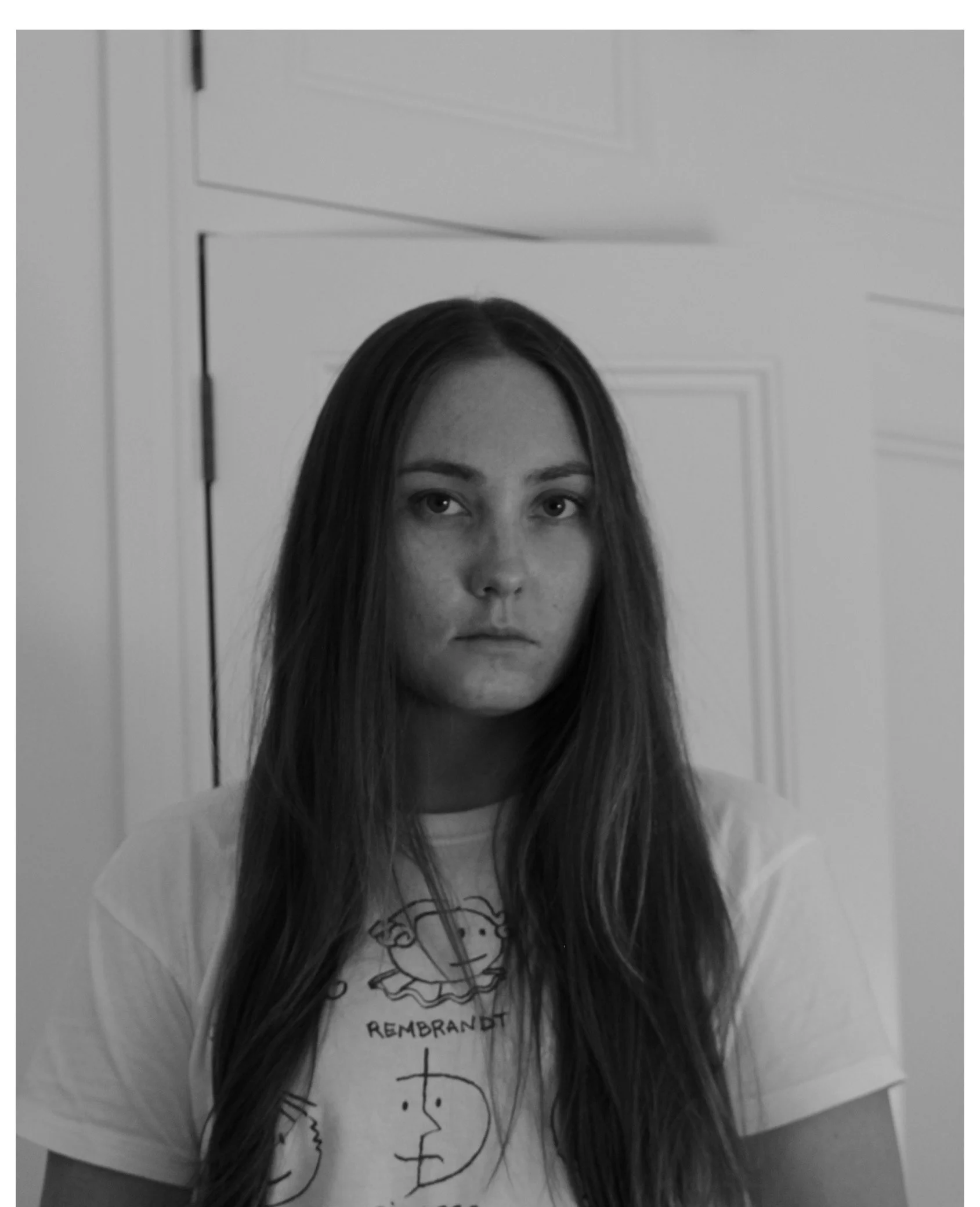 Black and white photo of a young woman with long hair, wearing a white t-shirt with cartoon faces and the text "Rembrandt." She is standing in front of a door with a panel design.