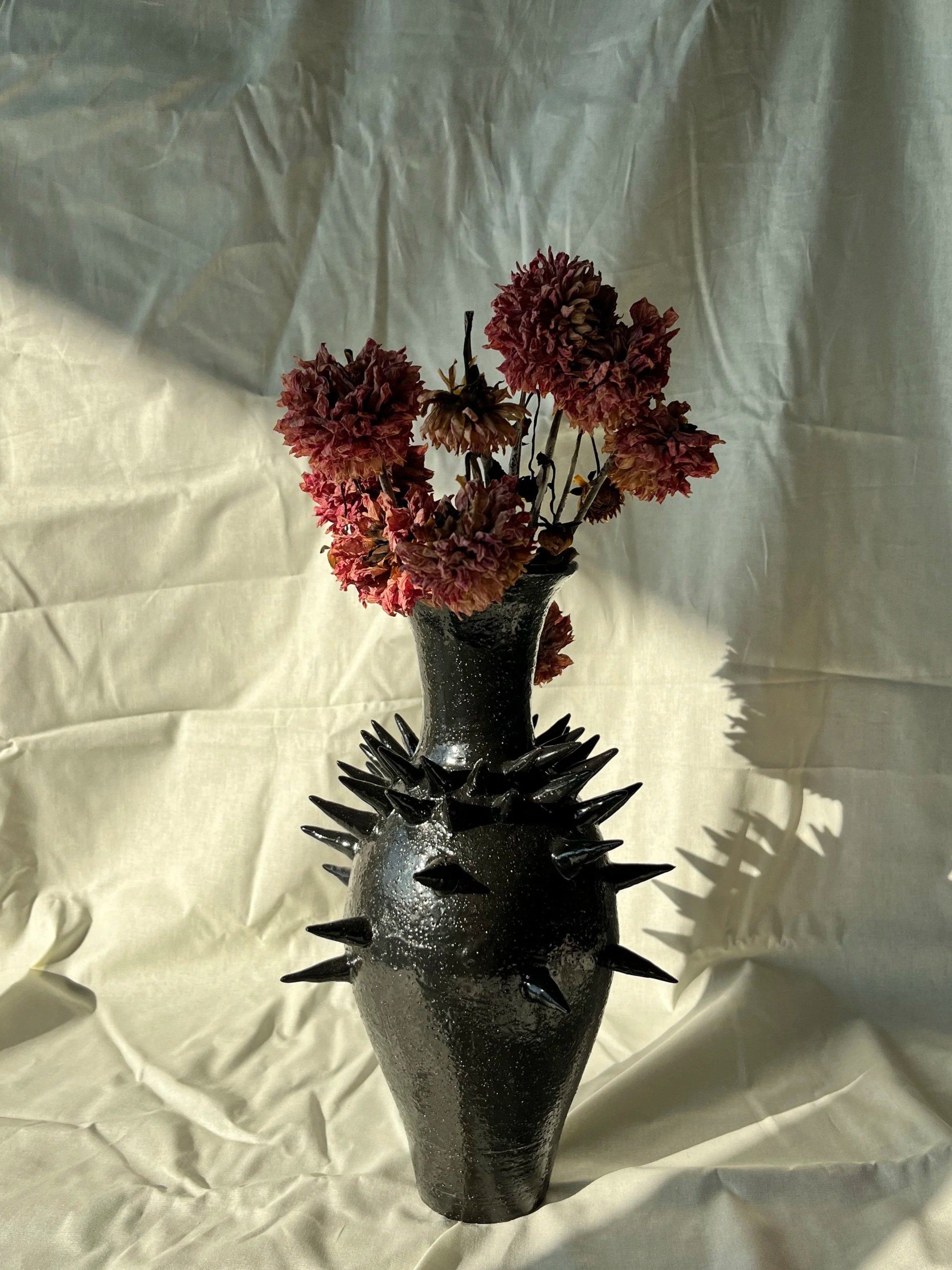 A spiked black ceramic vase with a textured surface holds dried pinkish-brown flowers, placed on a crinkled white cloth.
