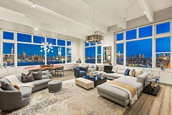 A modern living room with large windows showing a city skyline at night, furnished with a sectional sofa, armchairs, rugs, and a chandelier. Homeowner, condo, high-rise, assets, luxury.