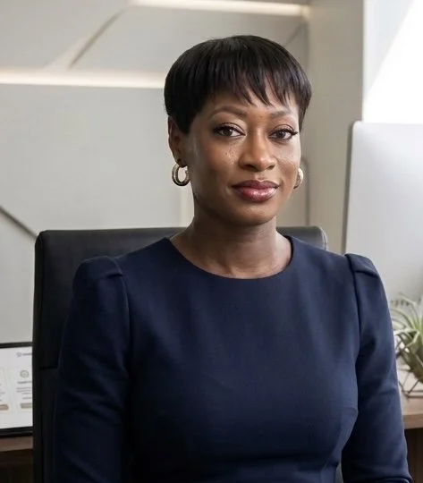 A woman with short black hair wearing a dark blue top, sitting in an office chair in front of a computer. Pixie cut, black woman, beautiful black woman, successful black woman, insurance agent, asset advisor, risk advisor, asset risk advisor.