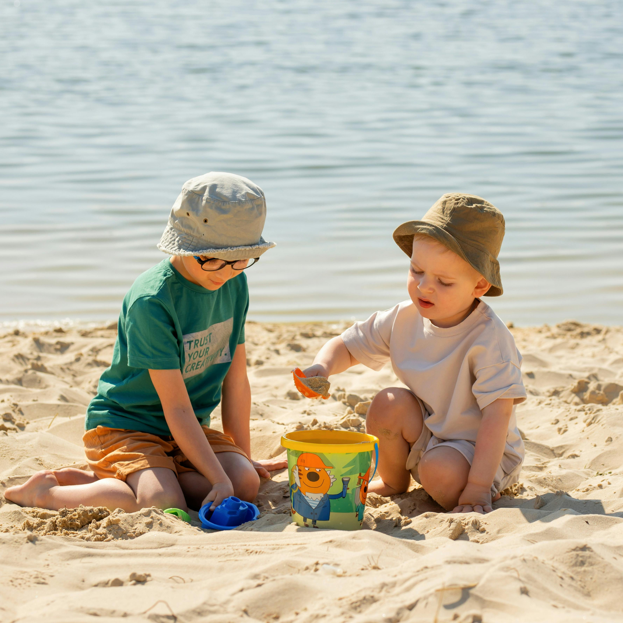 Dos niños adultos jugando en la arena en la playa, uno con gafas y sombrero gris, y otro con sombrero marrón claro, rodeados de juguetes de arena y un cubo con personajes animados de dibujos animados.