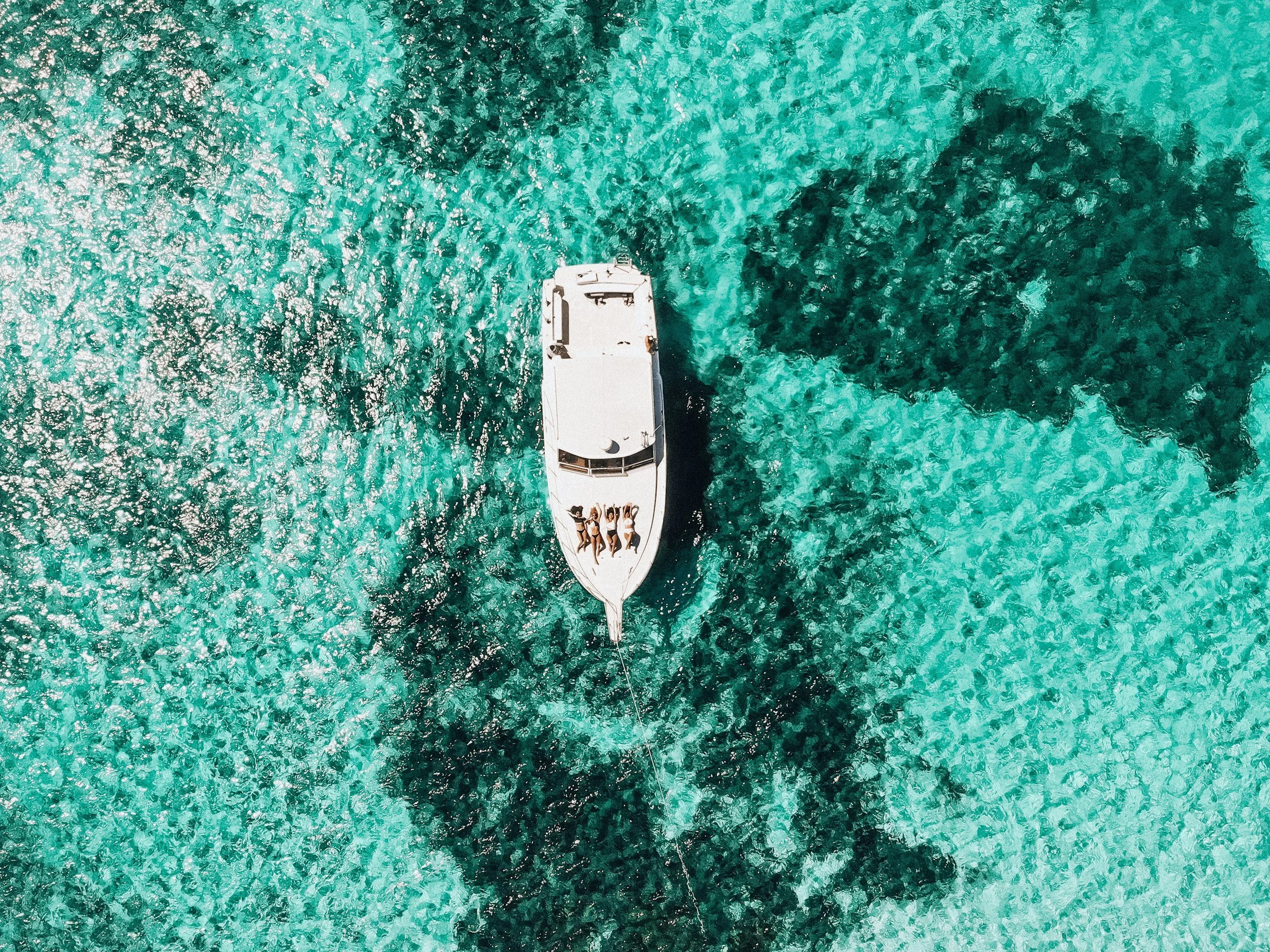 Imagen aérea de un yate navegando en aguas cristalinas con personas en la cubierta y arrecifes en el fondo.