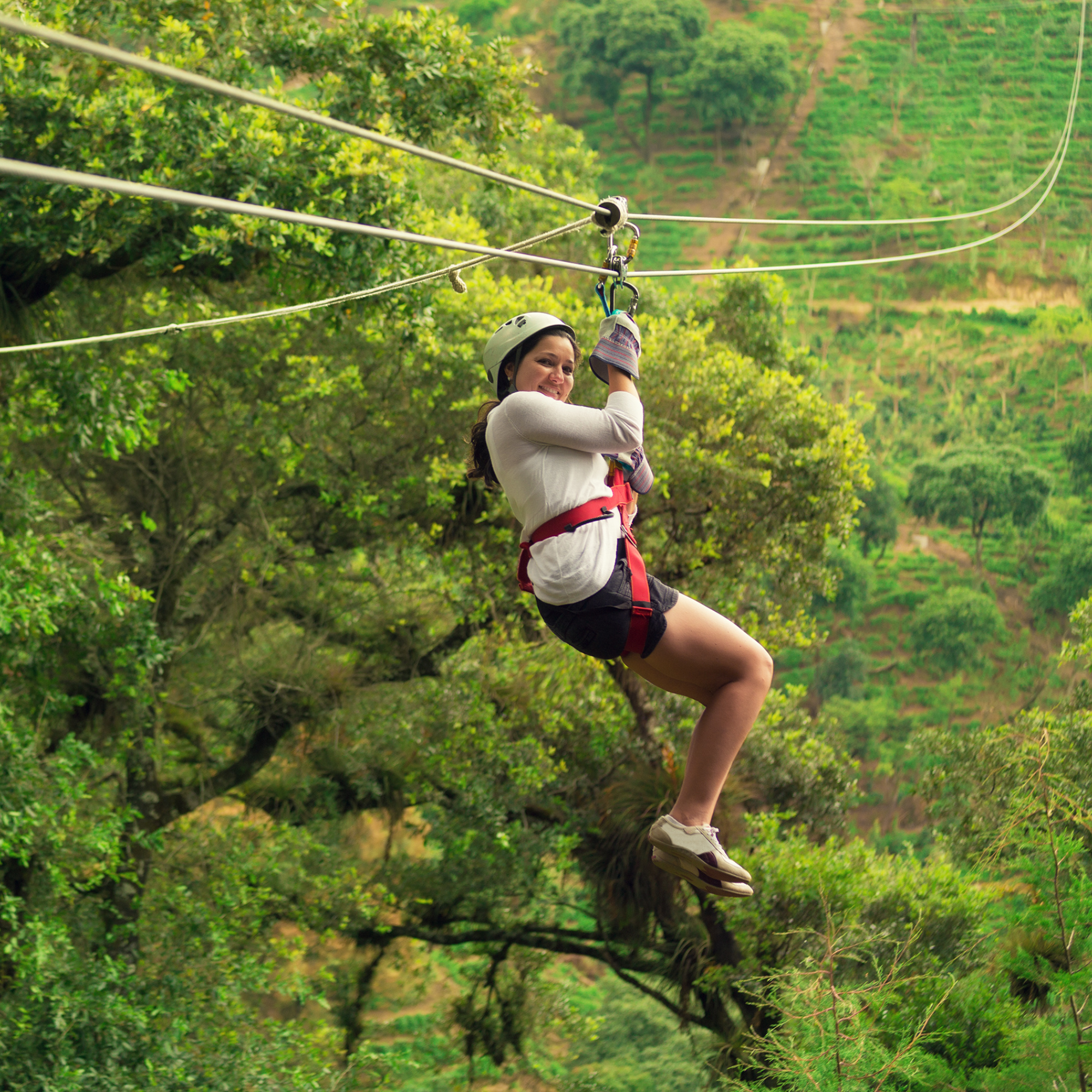 Mujer sonriendo en tirolesa en medio de un paisaje natural con árboles verdes y colinas.