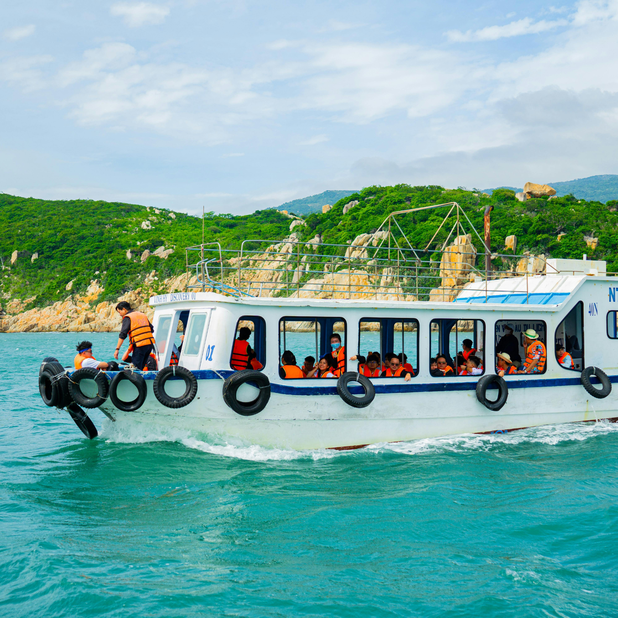 Ambulancia marítima con varias personas a bordo, algunas usando chalecos salvavidas, navegando en agua frente a una zona montañosa con vegetación y cielo parcialmente nublado.