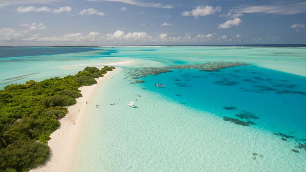 Vista aérea de playa con agua cristalina y arrecifes, bordeada por vegetación y cielo con nubes.