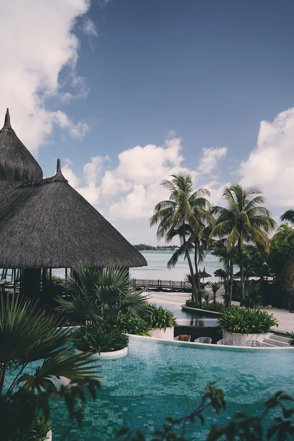 Paisaje tropical con palmeras, cabañas con techos de paja, piscina y vista al mar, cielo con nubes blancas y parcialmente nublado