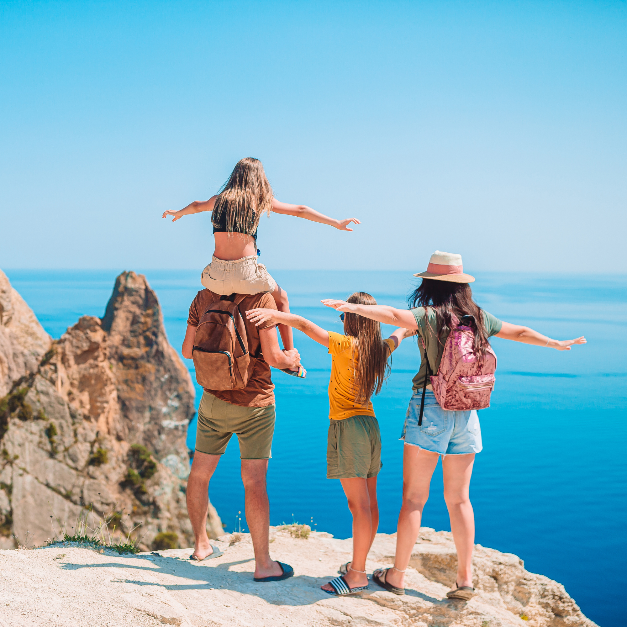 Personas caminando en un borde rocoso junto al mar en un día soleado, vista desde atrás, con una mujer con sombrero de paja y mochila y otras personas en grupo disfrutando del paisaje.