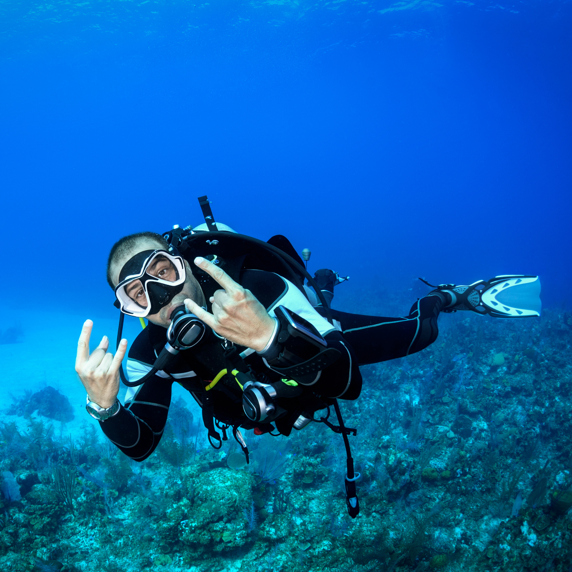 Persona practicando buceo en el océano con equipo y máscara de protección, haciendo señal de paz con la mano, fondo de arrecife de coral y agua azul clara.