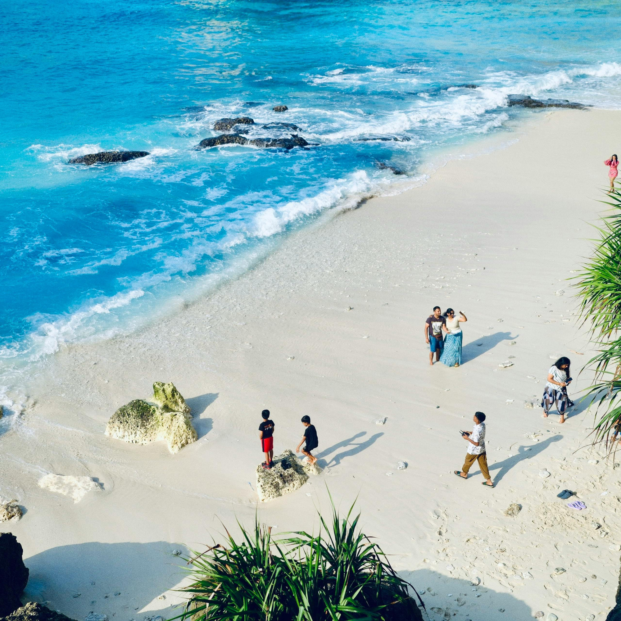 Playa de arena blanca con rocas en el agua azul y personas paseando y tomando fotos, vegetación en primer plano, cielo despejado.