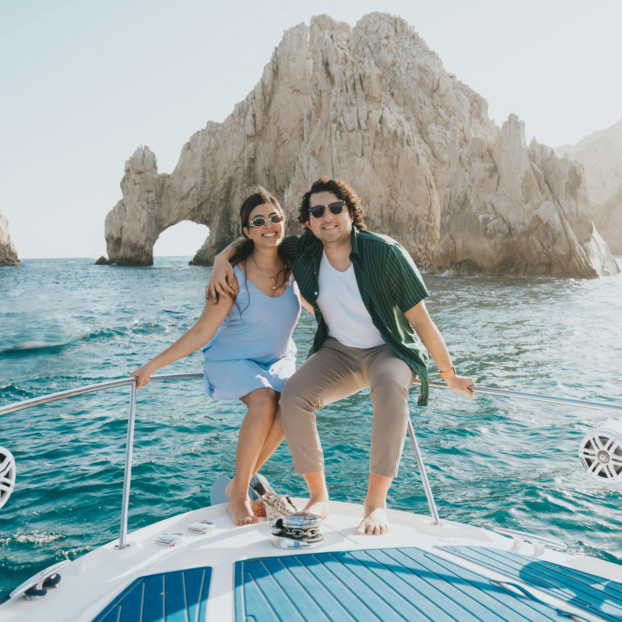 Pareja sonriendo en un yate frente a un arco de piedra en el mar