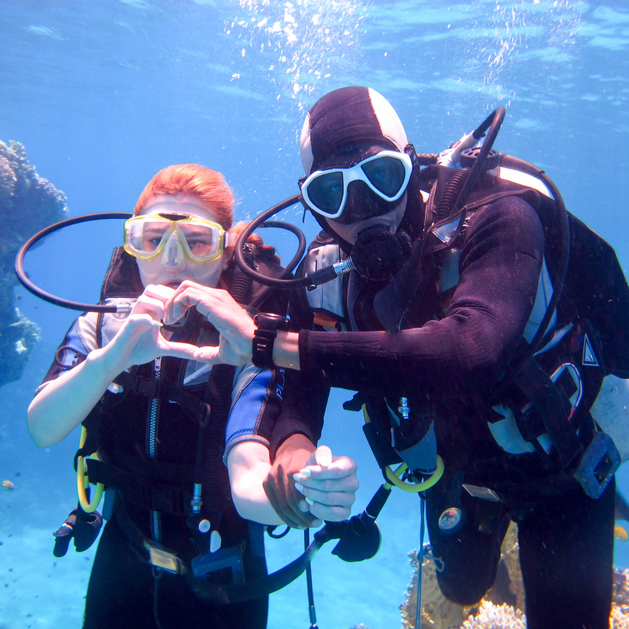 Dos personas practicando buceo en el océano, usando trajes de buceo, máscaras y tanques de oxígeno, rodeadas de agua y coral.