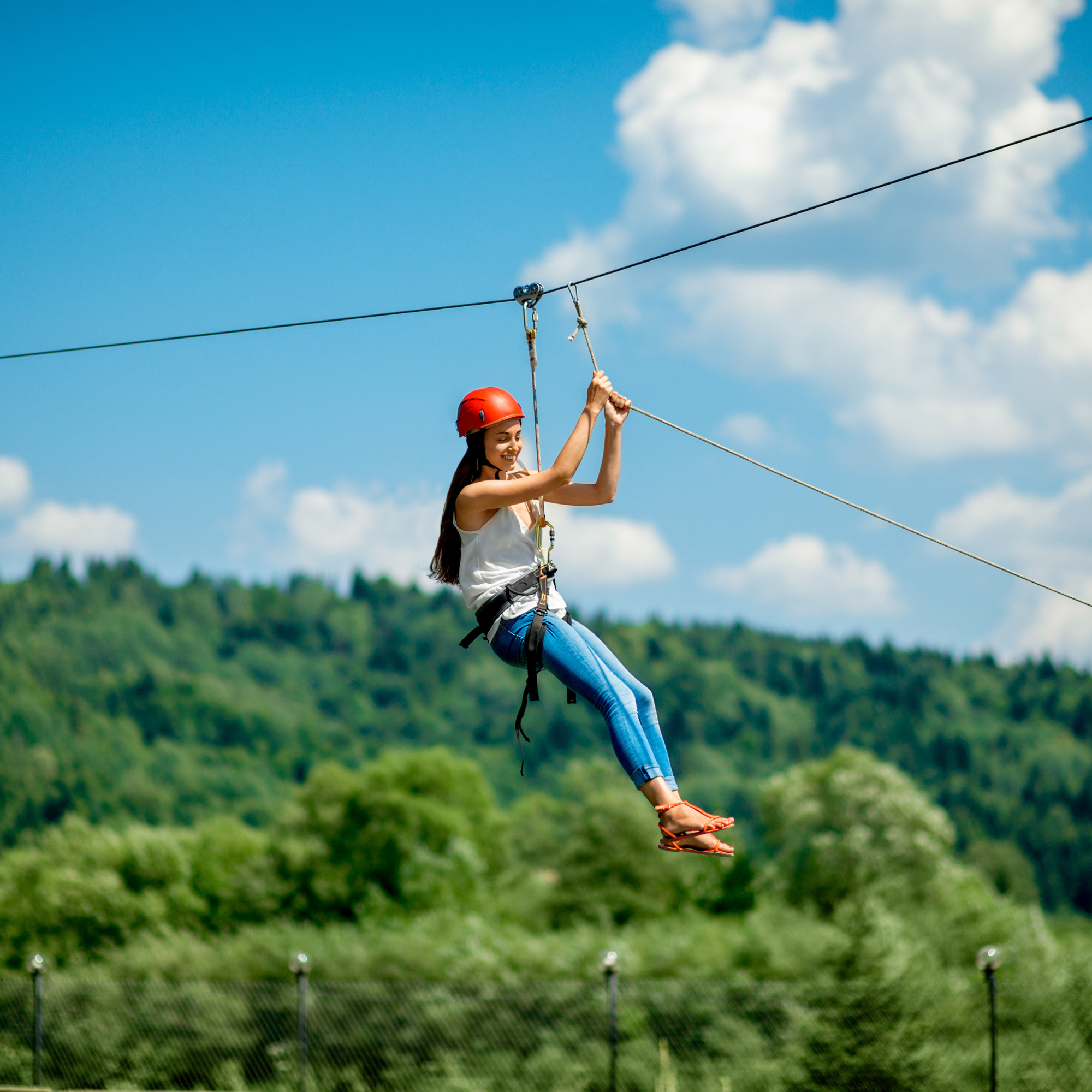 Joven mujer en arnés y casco disfrutando de una actividad de tirolesa en un entorno natural con césped y árboles verdes, cielo despejado con algunas nubes.