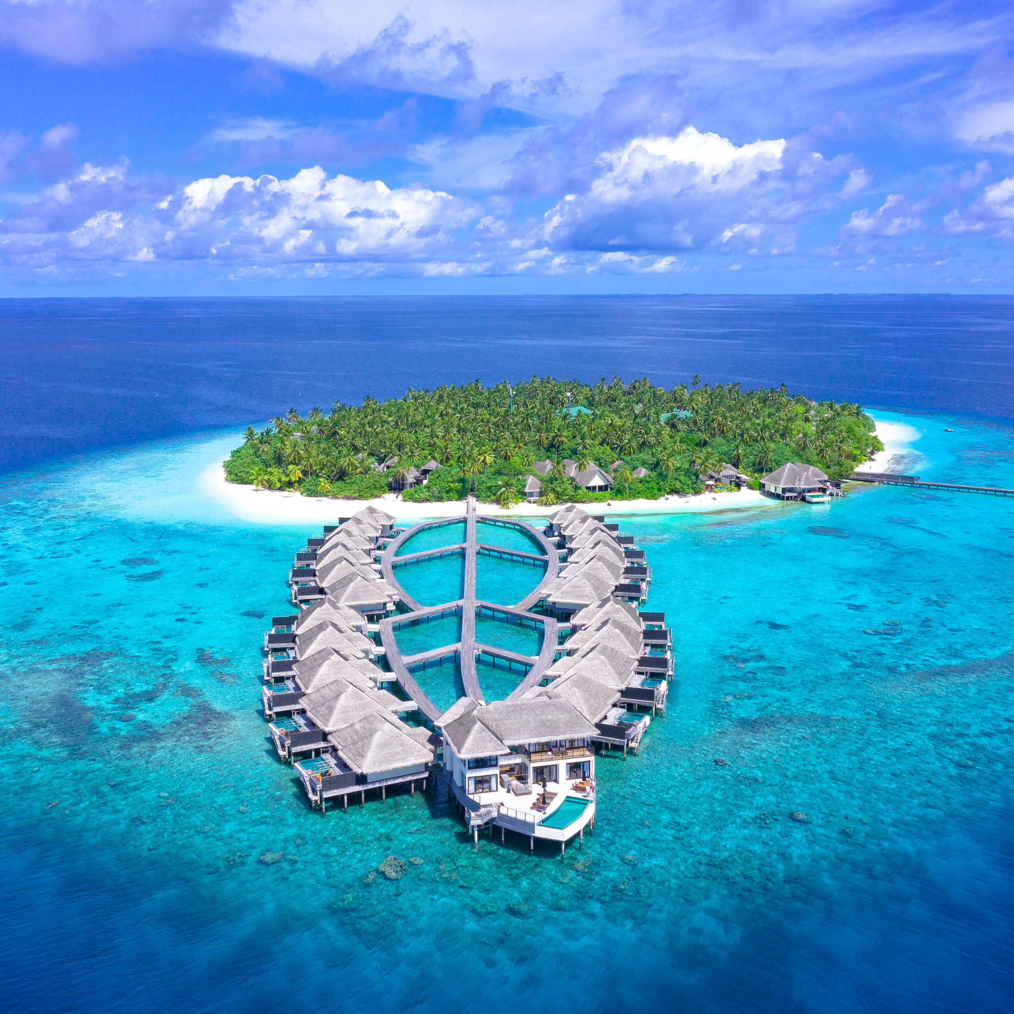 Un complejo turístico sobre el mar con villas al borde del agua, rodeado de aguas azules y verdes, con una piscina y vegetación tropical en primer plano, cielo parcialmente nublado.